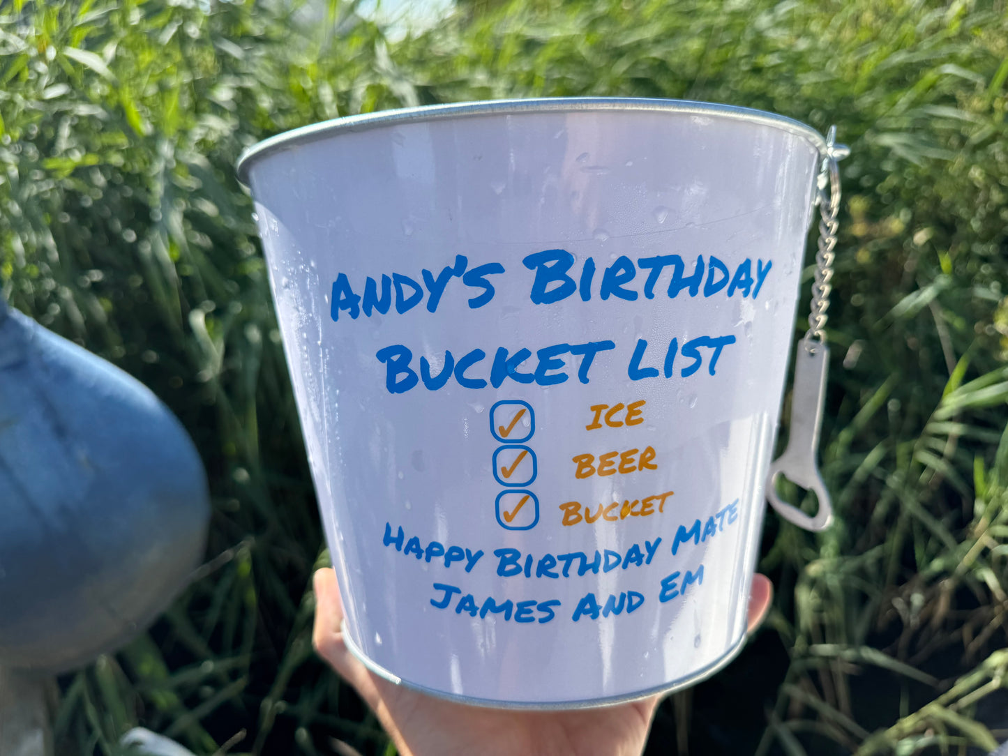 Personalised Beer gift bucket, with the recipients name written on it and a personal message, the bucket is being held up with long grass in the background