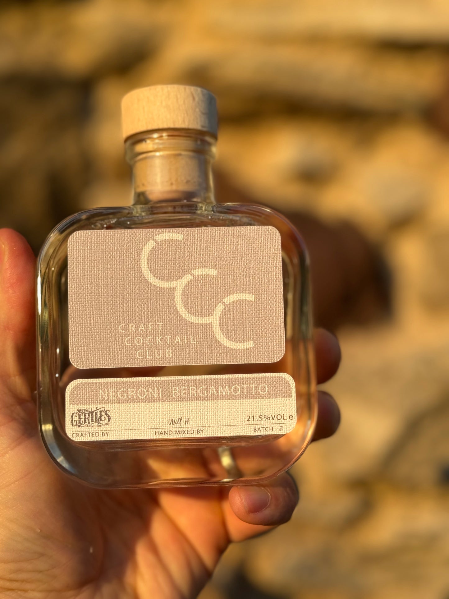 pictrue of a white negroni in a clear glass bottle being help up in a mans hand with the sun setting behind