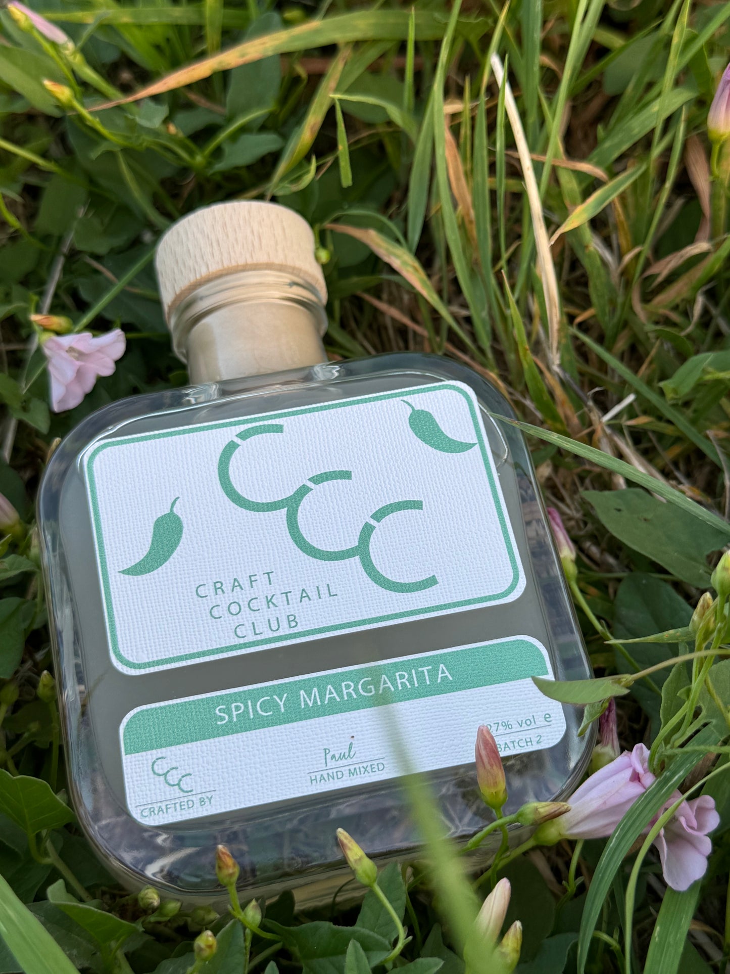 picture of a square cocktail bottle containing spicy margarita, lying flat in some green grass and pink flowers