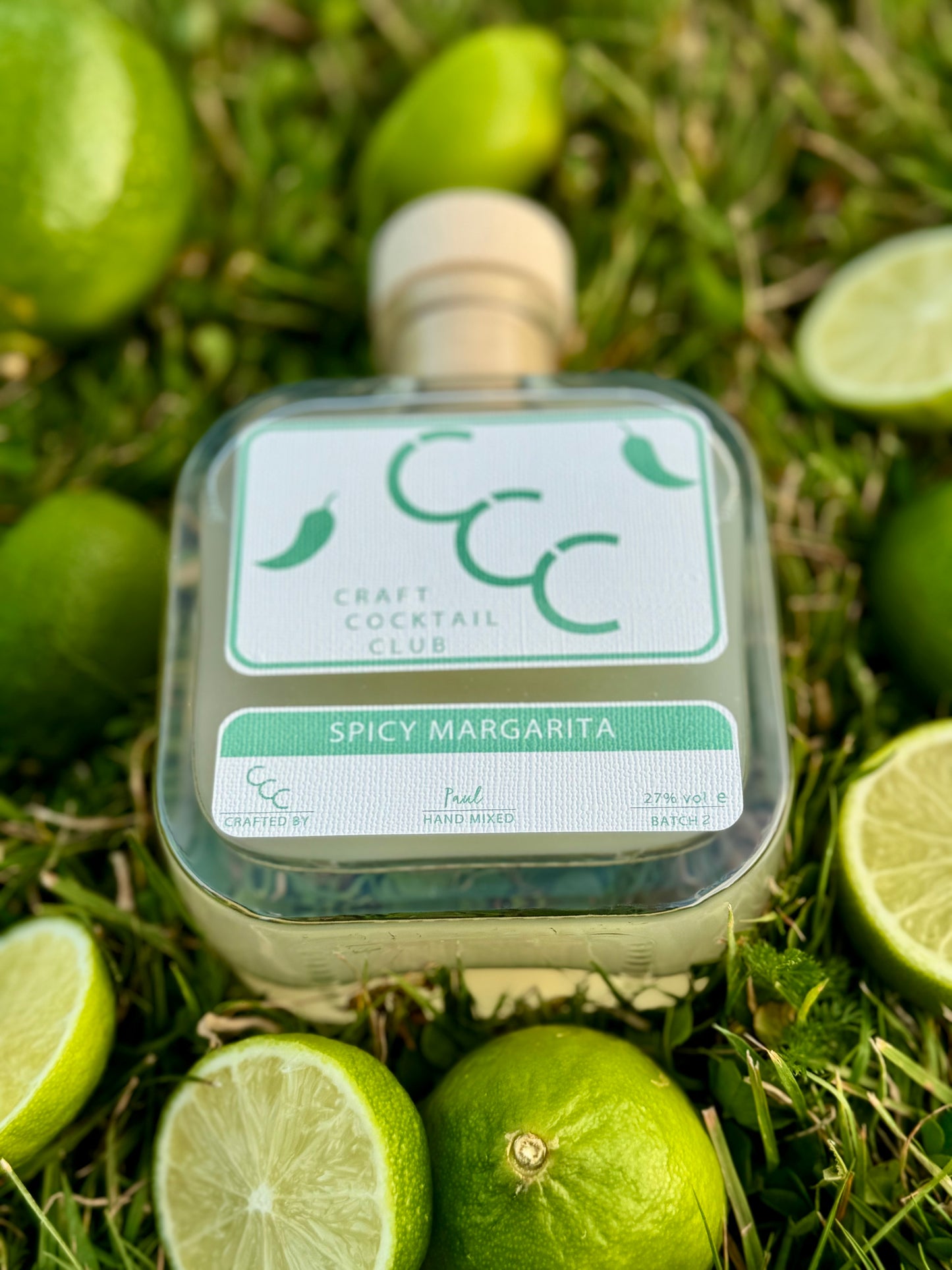 Picture of a bottle of craft cocktail club spicy margarita, lying flat on green grass surrounded by fresh cut limes