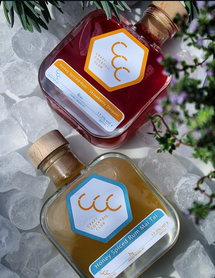 Picture of two bottles of ready to drink cocktails surrounded by ice cubes with foliage blurred on the edge of the picture, labels on the bottles say Craft Cocktail Club