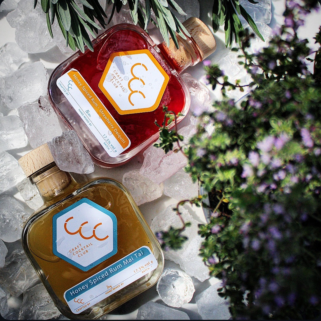 Picture of two bottles of ready to drink cocktails surrounded by ice cubes with foliage blurred on the edge of the picture, labels on the bottles say Craft Cocktail Club