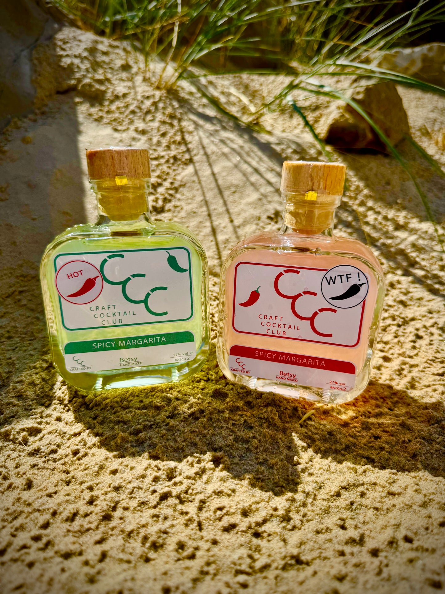Two small bottles of spicy margarita with chili peppers on a beach, surrounded by fresh limes with a gift box in the background - Label reads Craft Cocktail Club Spicy Margarita