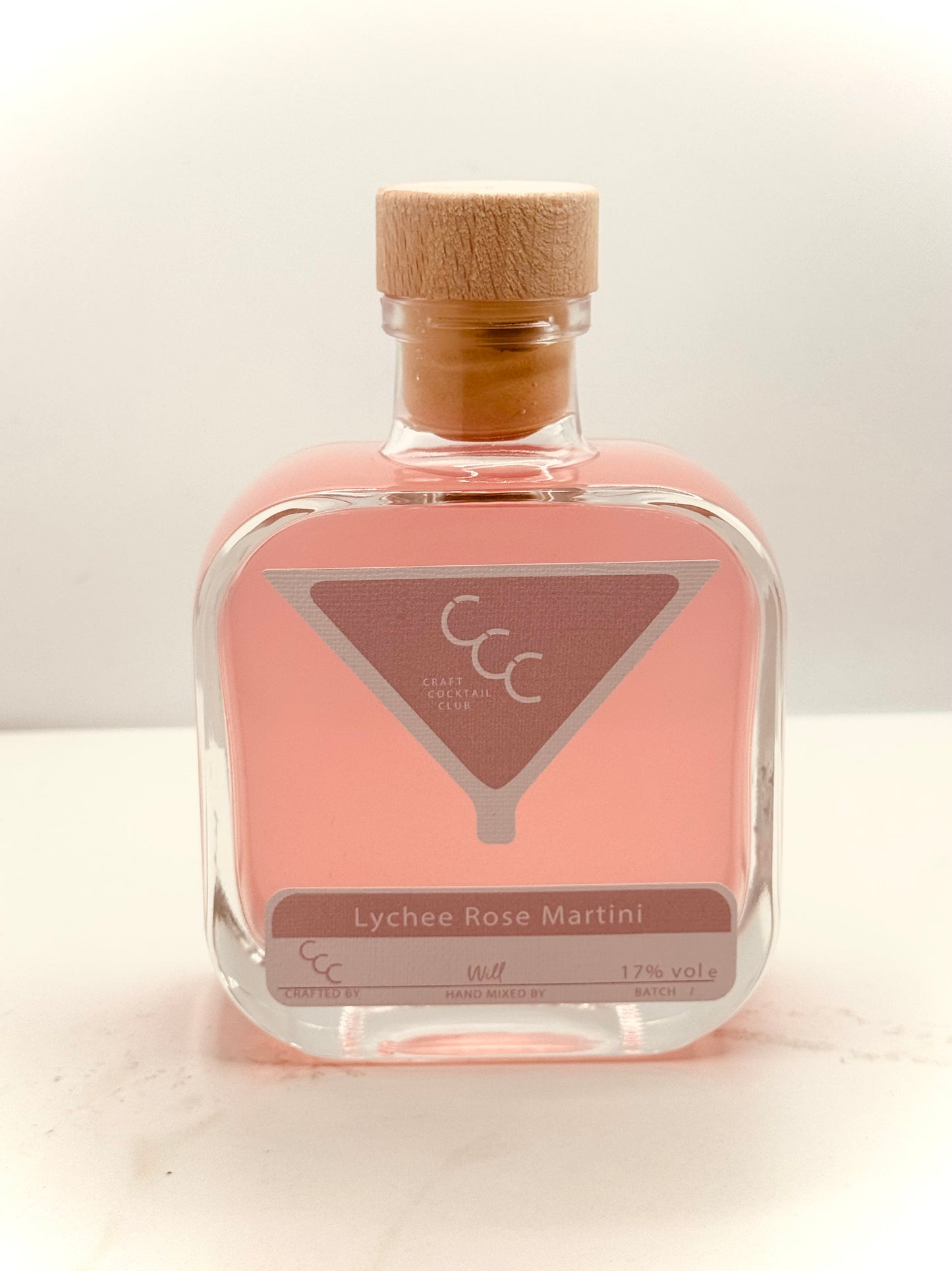 Picture of a pink Square glass bottles of Cocktail, on a marble surface. Label reads Craft Cocktail Club. Lychee rose martini