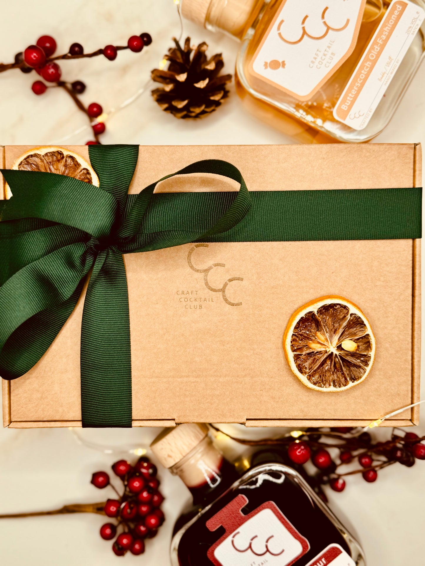 A picture of a Craft cocktail club gift box, wrapped in a red ribbon surrounded by winter berries and bottles of ready to drink cocktails