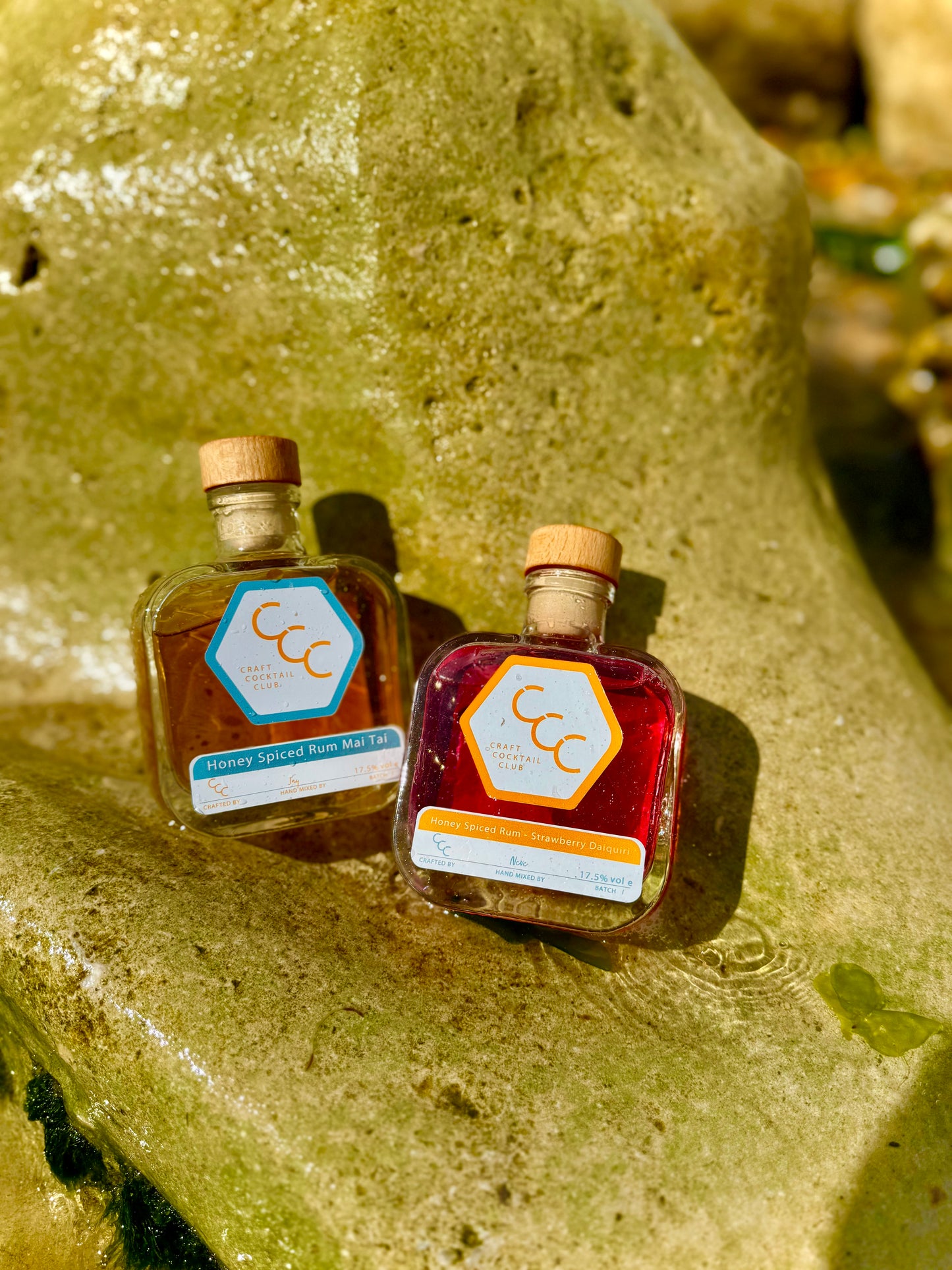 Picture of two bottles of rum cocktails sat on a rock with water splashing up on them.