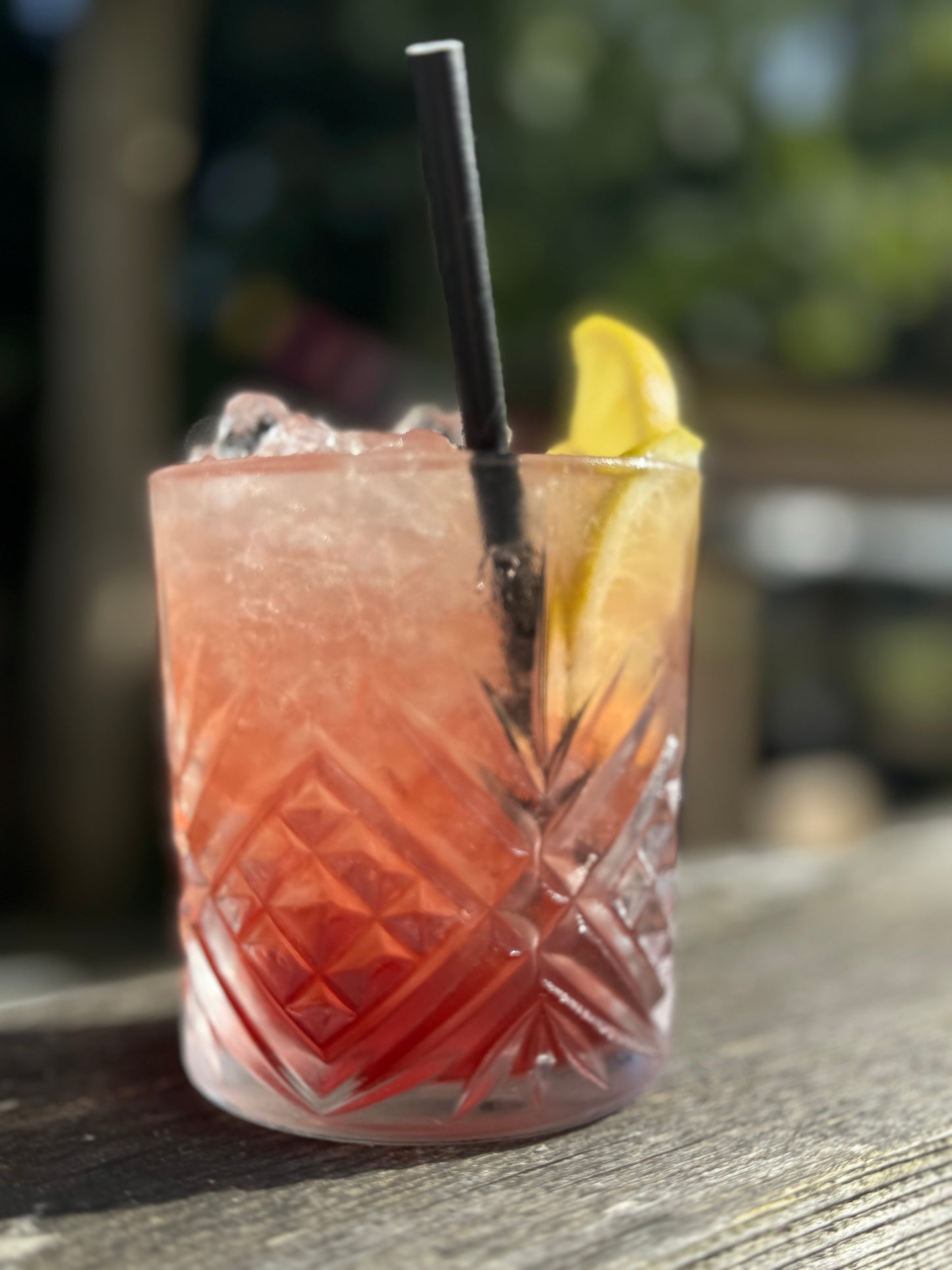 single glass of a Bramble cocktail outside on an oak table with a straw and lemon