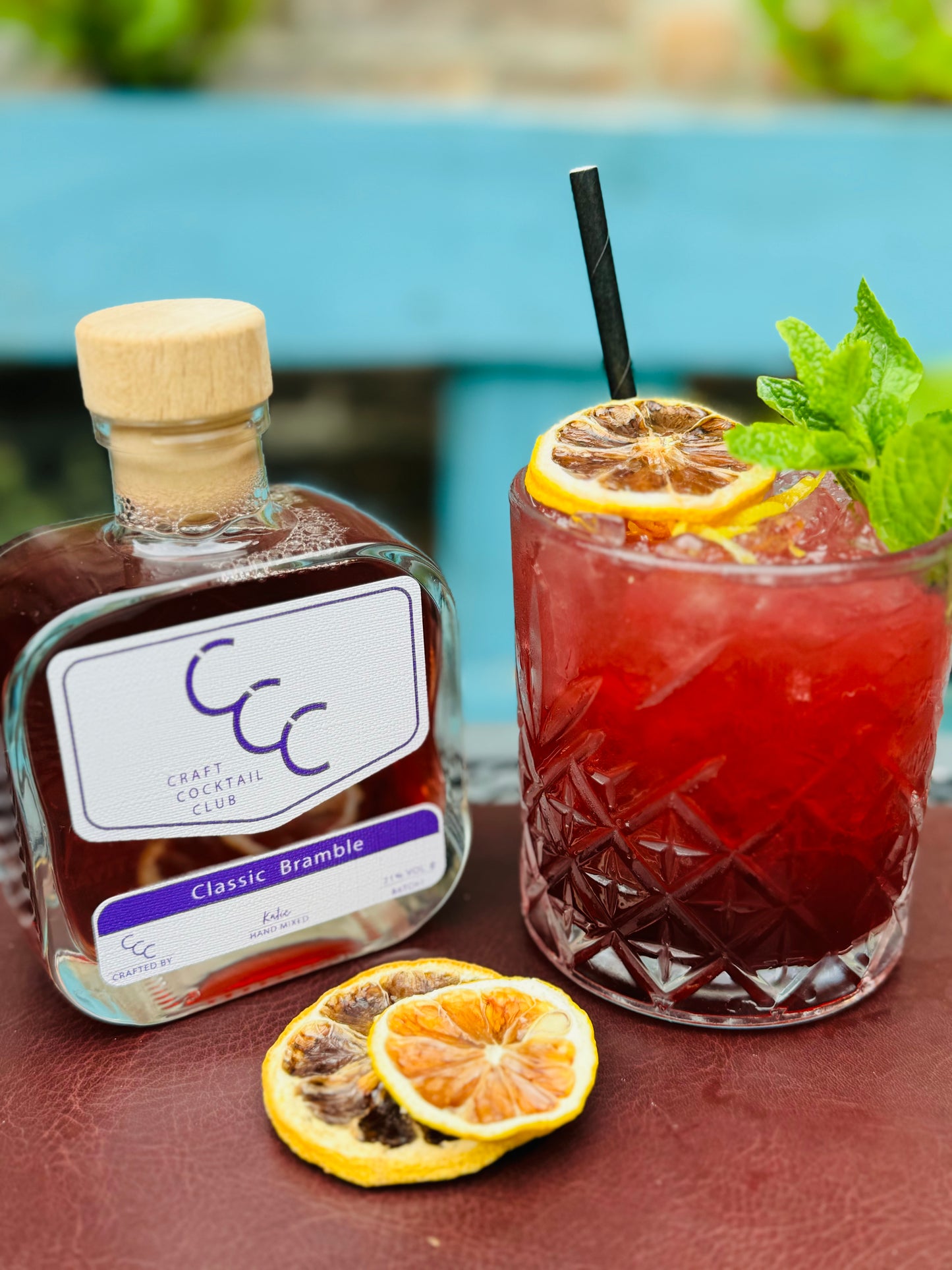 picture of a bramble craft cocktail club bottle and purple drink outside on a sunny day with a lemon garnish