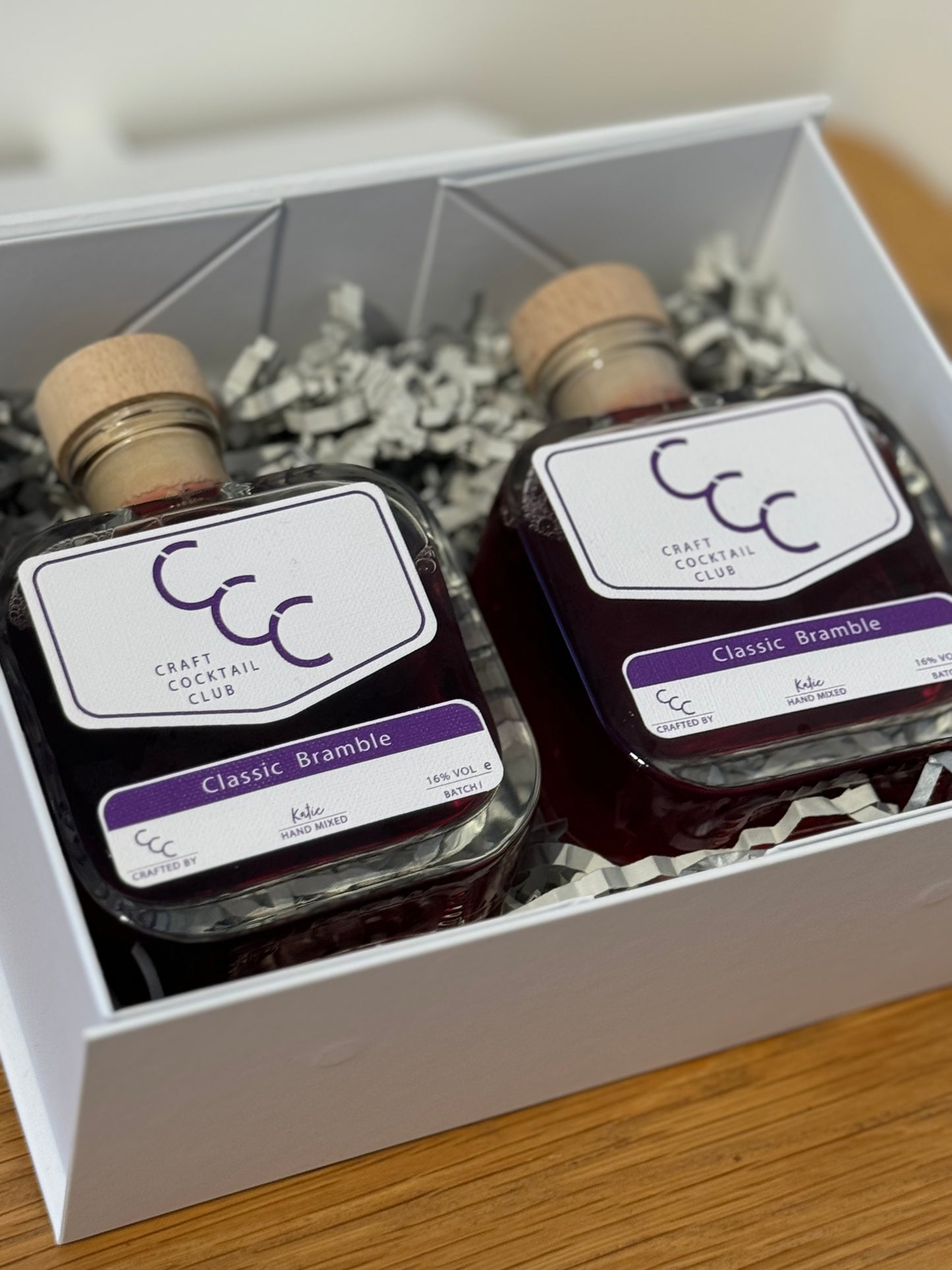 Picture of two square glass bottles filled with purple liquid, sat together in a gift set box on and oak table bottles labels say Classic Bramble Craft Cocktail club