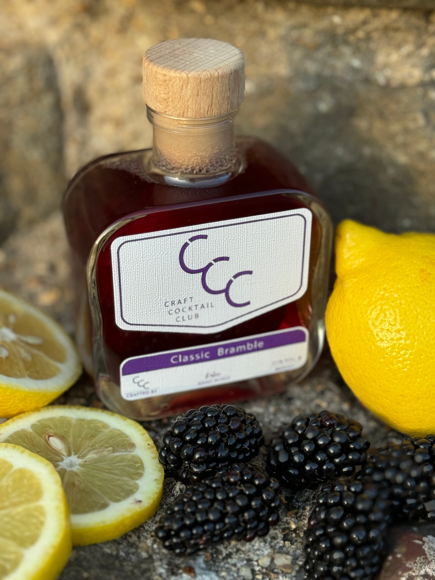 Picture of a Glass bottle with deep purple liquid inside surrounded by fresh blackberries and cut lemons, label reads craft cocktail club. Classic bramble