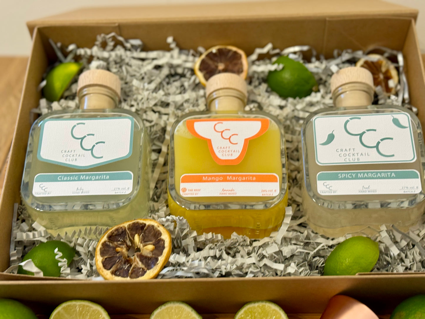 picture of a gift box with three margaritas inside, the bottles a green and orange and. the box is surrounded by green fruit
