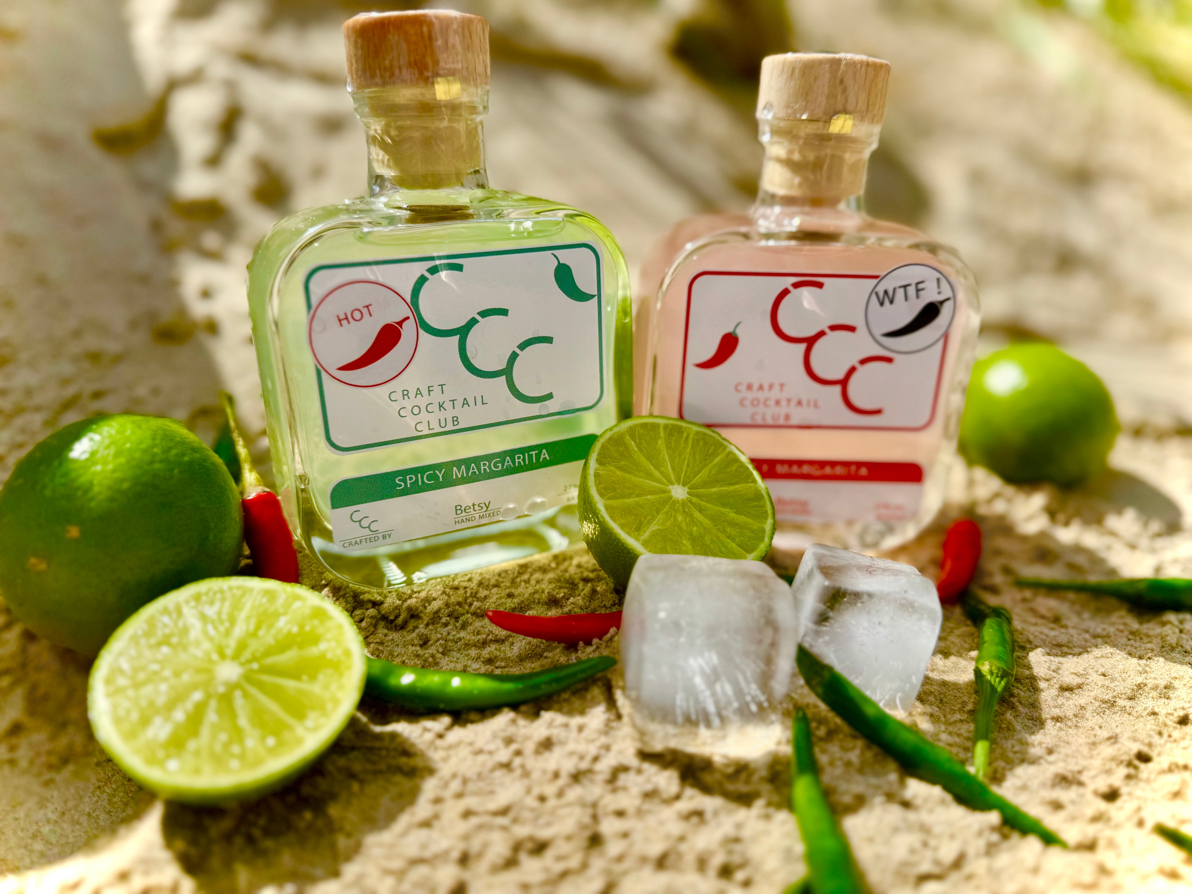 Picture of two bootle of cocktail in a square glass bottles, one has light green liquid the other light pink. they are sat on a beac surrounded by limes and chillies. the table reads Craft Cocktail Club- Spicy Margarita