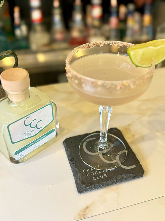 picture of a margarita glass with a lime on the edge on a coaster next to a pre made margarita bottle
