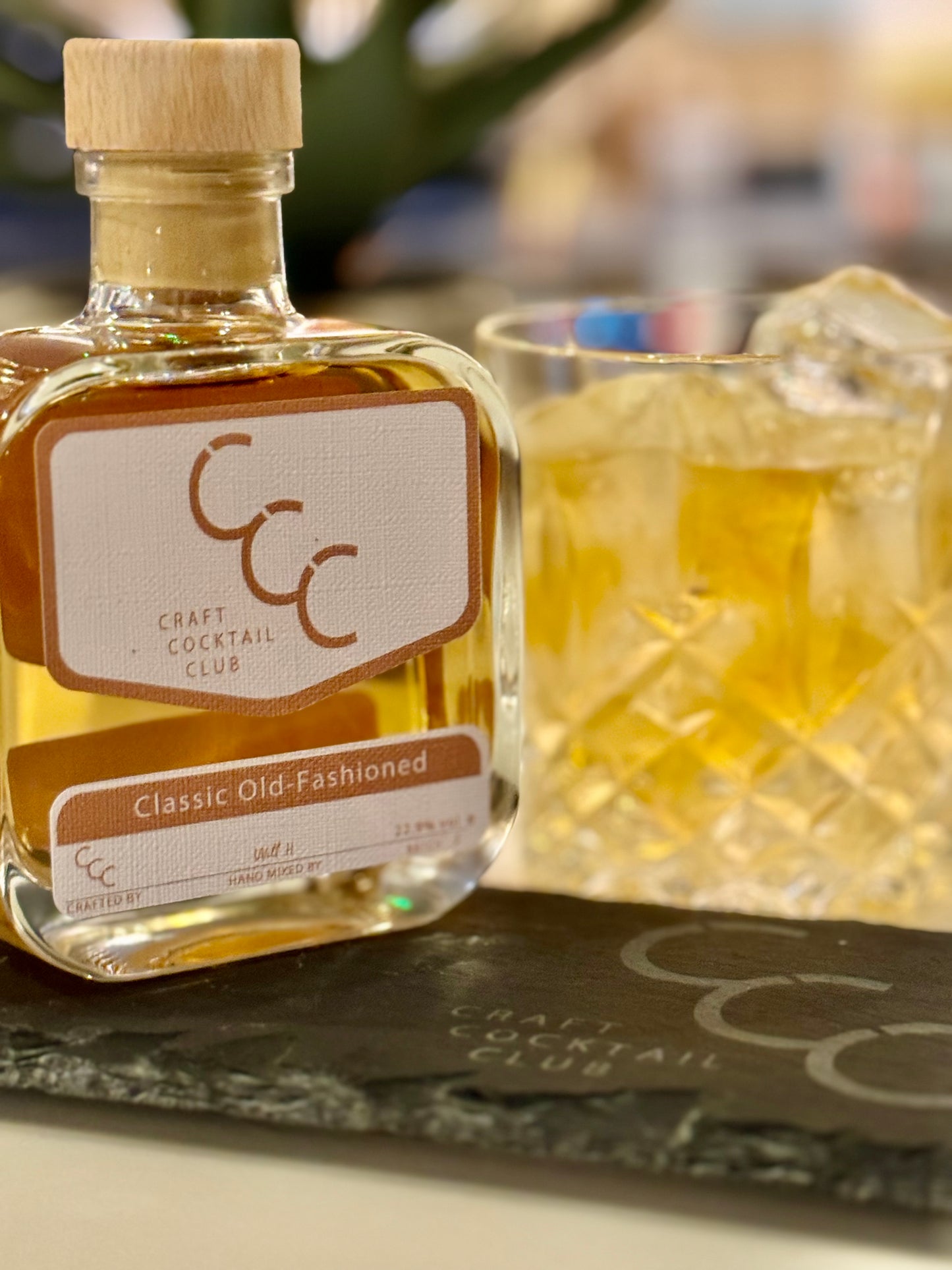 A picture of an Old Fashioned Cocktail in a crystal tumbler glass full of ice and golden liquid. Next to it is a ready mad bottle of the cocktail with a label that reads Old Fashioned - Craft cocktail Club
