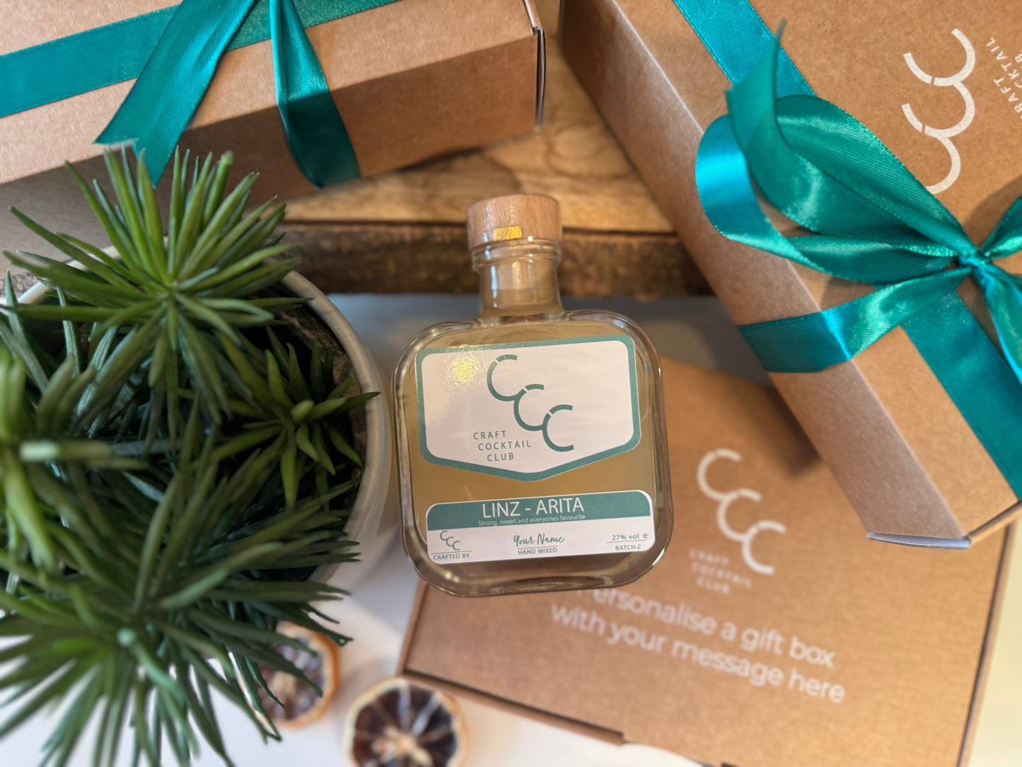 Cocktail bottle with a plant and gift box on a wooden surface - The Square glass bottle contains lime greed coloured Margarita, and the label has been personalised to read Linz-Aritia