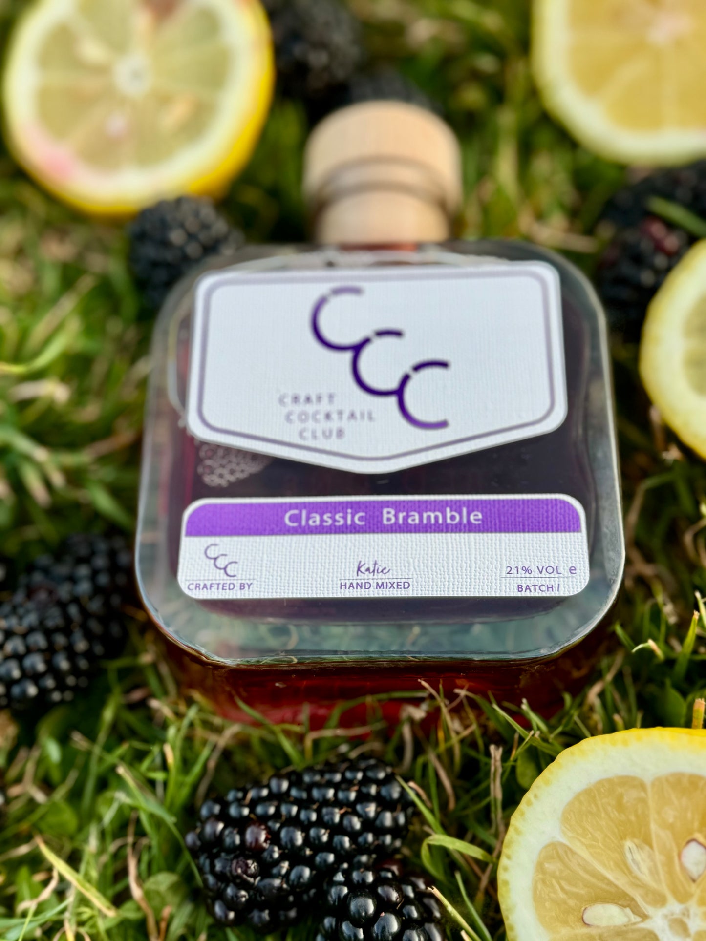 picture of a bramble cocktail from the craft cocktail club, phot has a slight blur and the bottle is flat on the grass surround by fresh blackberries and lemons