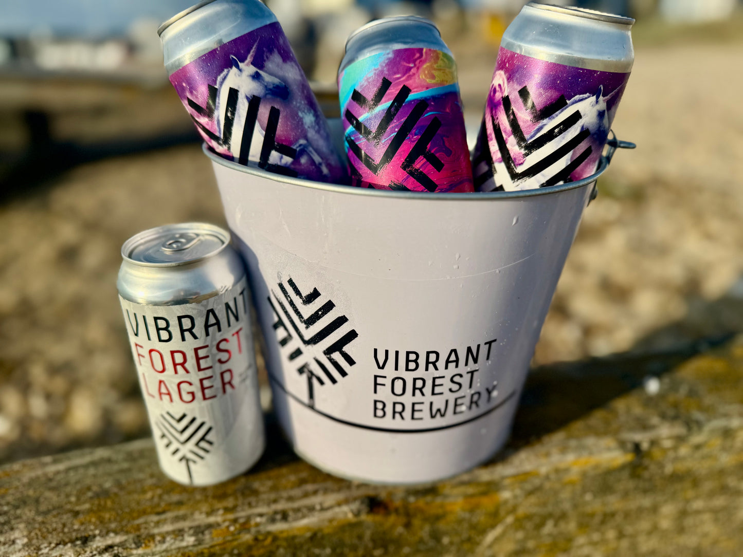Colorful beer cans in a bucket with 'Vibrant Forest Brewery' branding on a wooden surface.