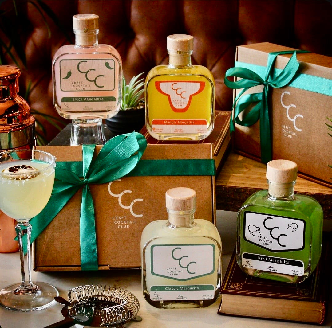 Craft Cocktail Club bottles with packaging and a cocktail on a table. Surrounded by gift boxes and cocktail accesories 