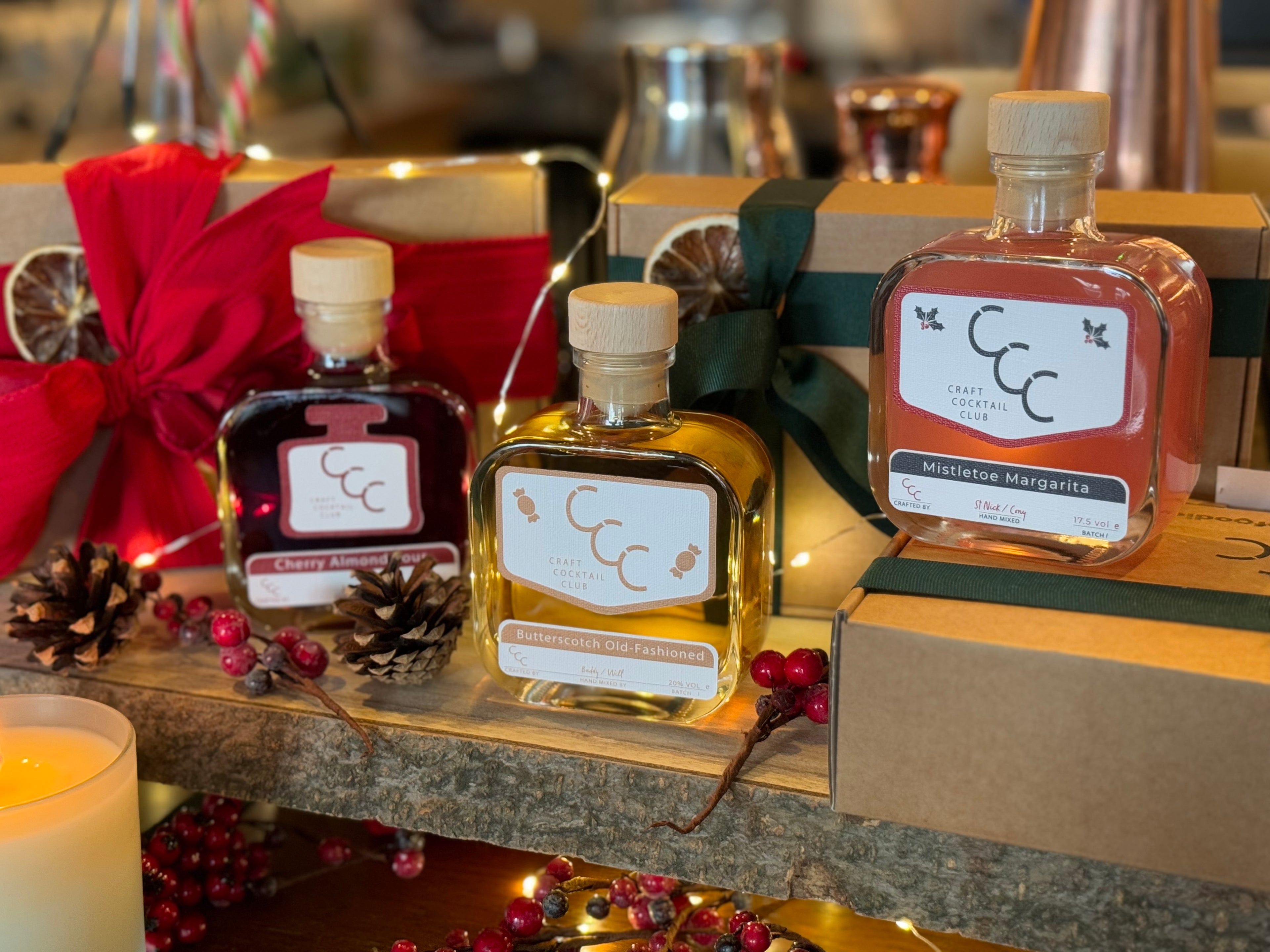 Picture of three small square glass bottles, next to a gift box. the bottle each contain handmade cocktails, In Dark Ruby Red, Whisky brown and a light pink. The labels read Cherry almond Sour - Old Fashioned and Mistletoe margarita. In the background are fairly lights and red Christmas berries 