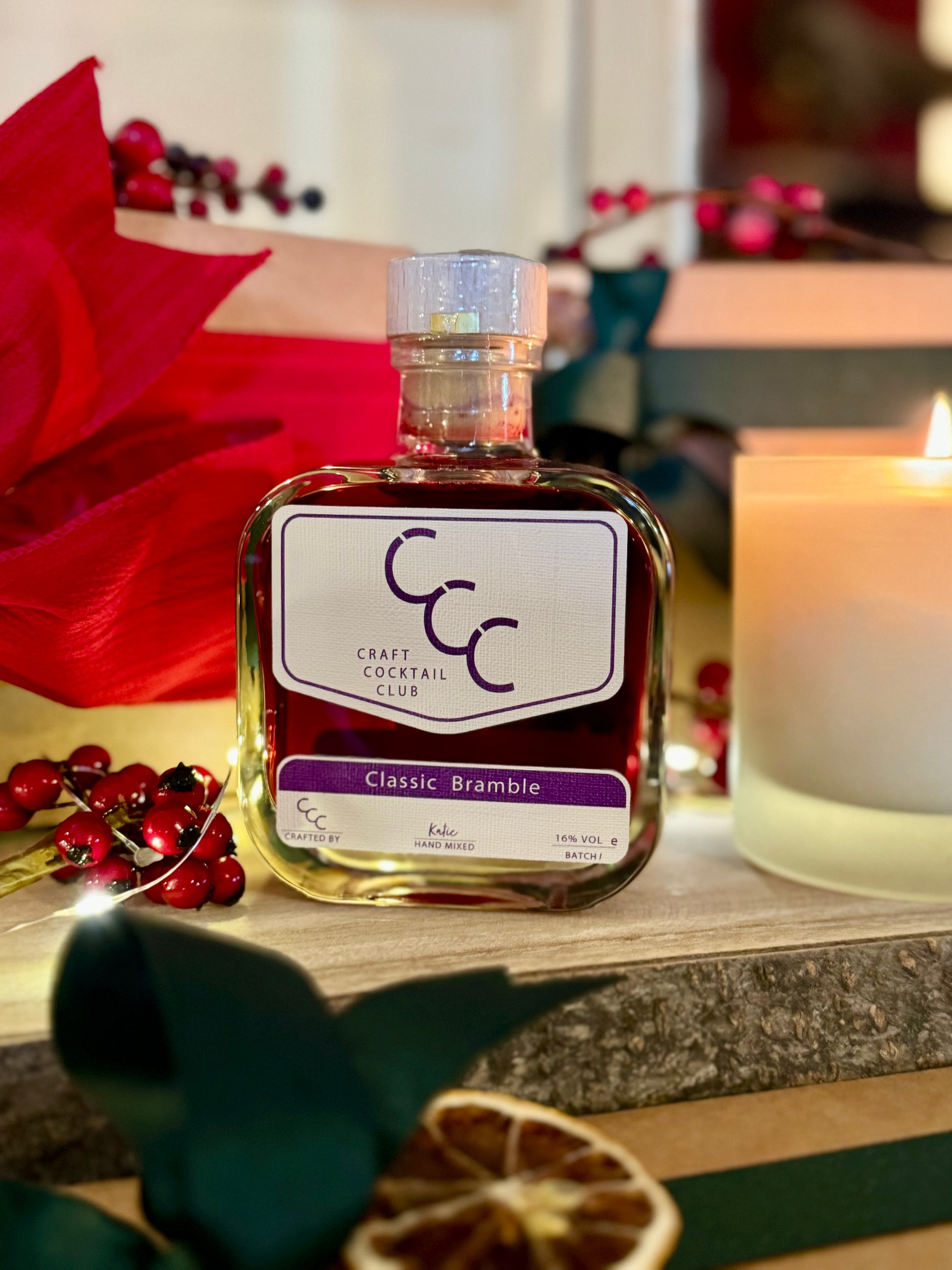 Picture of a square bottle of Bramble cocktail from Craft Cocktail Club, its sat on a rough wood board, with lights and candles in frame