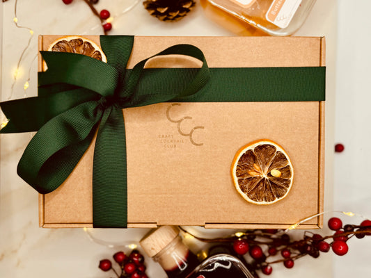 A festive gift box with craft cocktail club written on it, Wrapped in a green ribbon and surrounded by fruit and winter berries 