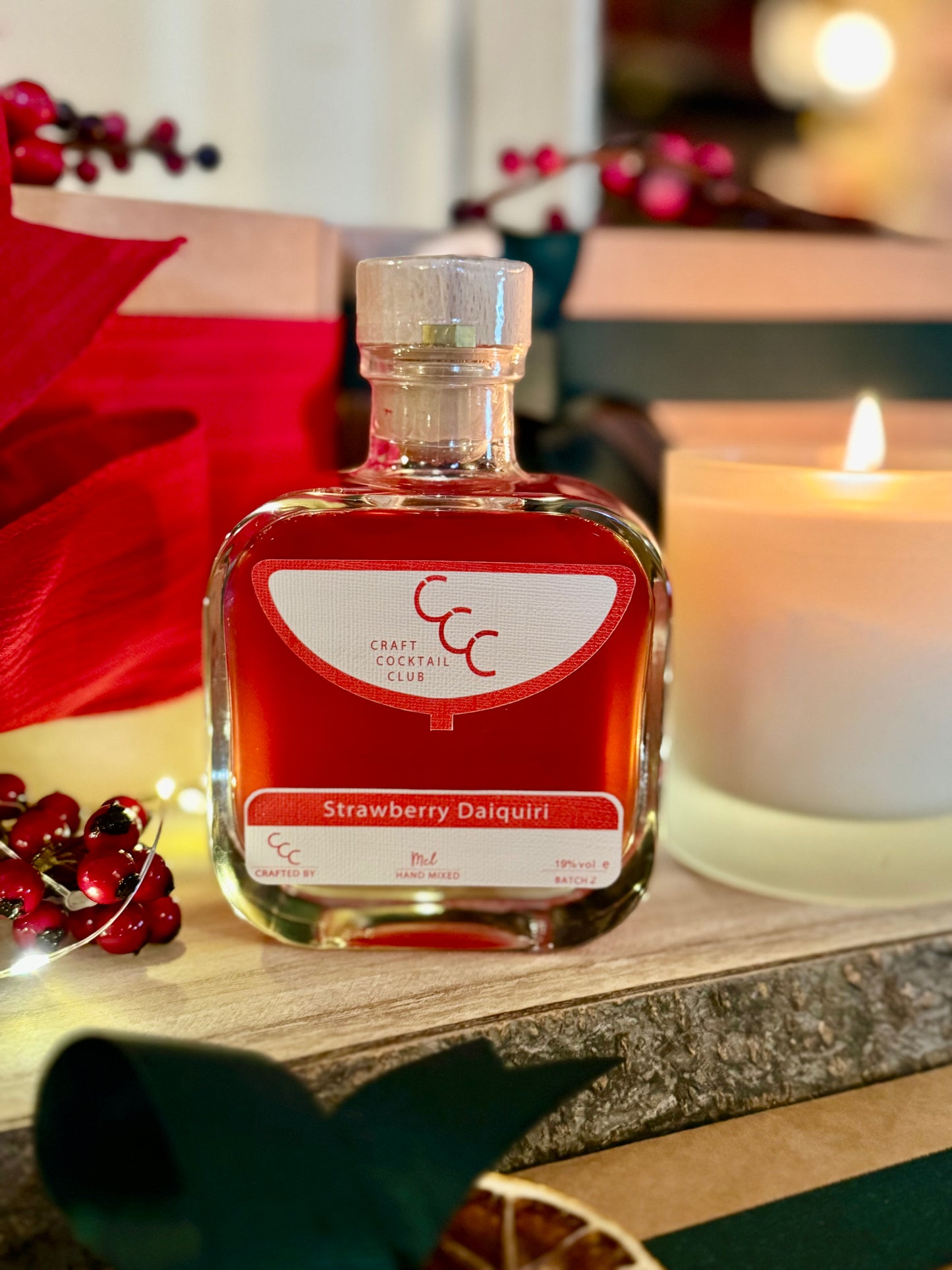 Picture of a luxury square glass bottle from Craft Cocktail Club, the bottle is on a rough wood board surrounded by candles and red berries. On the label it reads Strawberry Daiquiri 