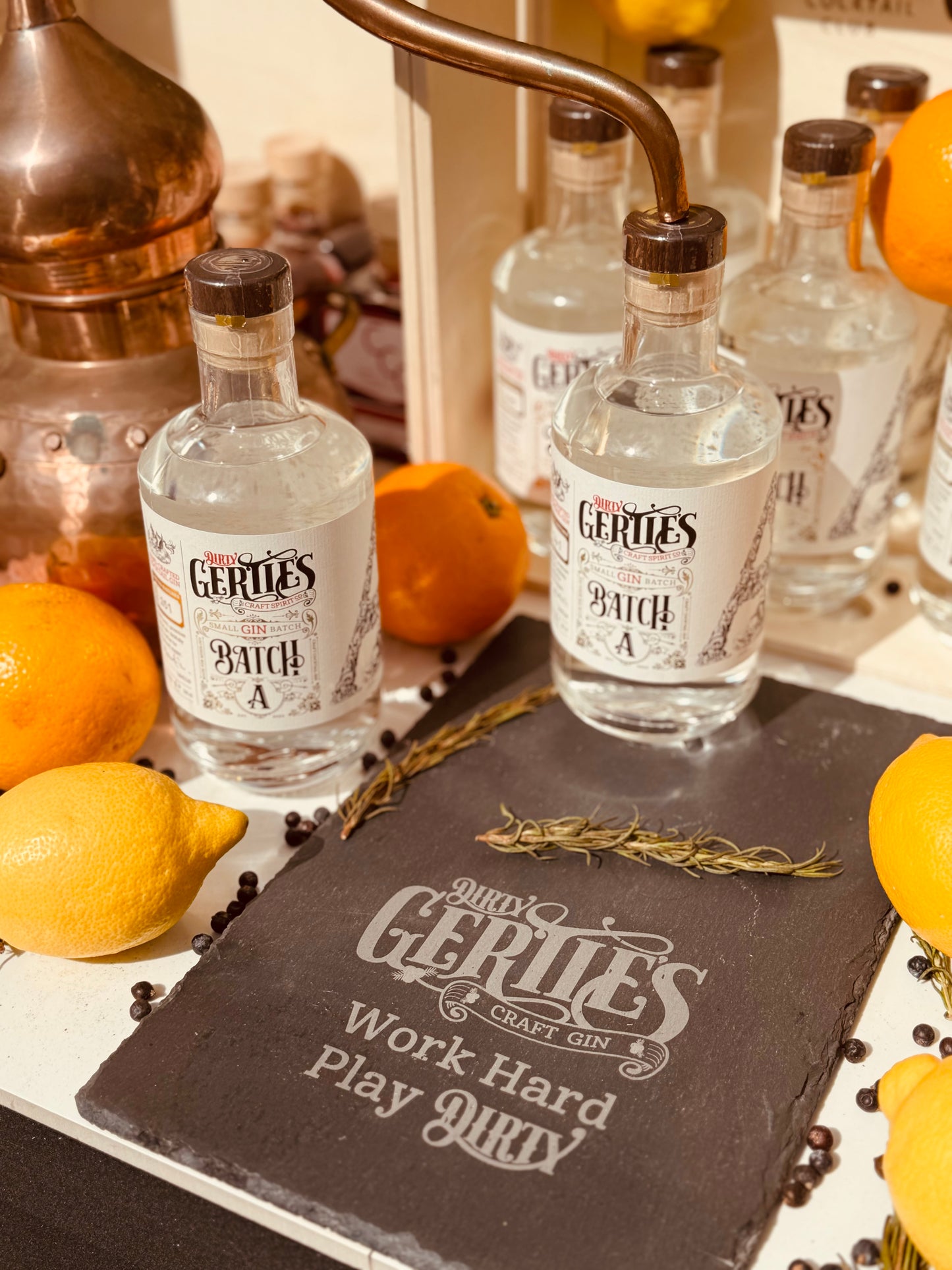picture of Dirty Gertie's gin bottle next to a small copper still with a slate with the words work har play dirty next to it 