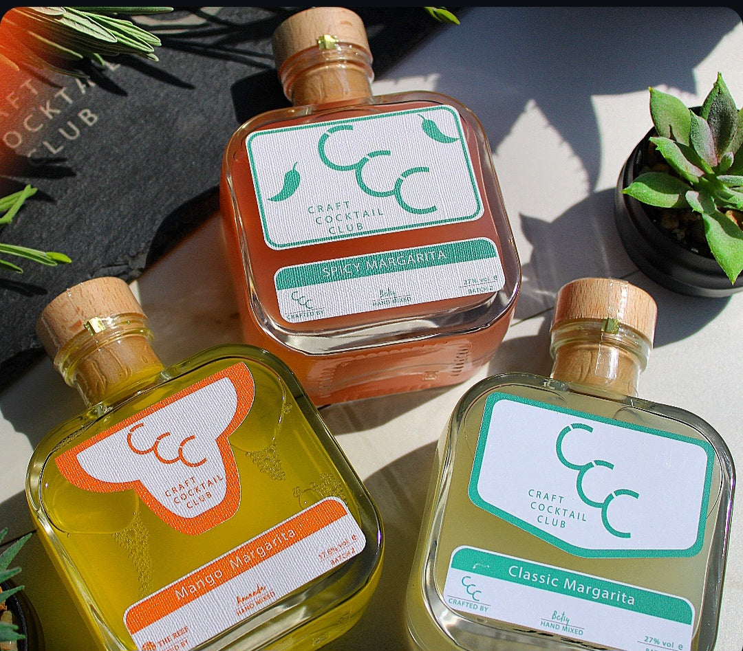 Three bottles of Craft Cocktail Club margarita mix on a surface with plants and a slate board in the background.