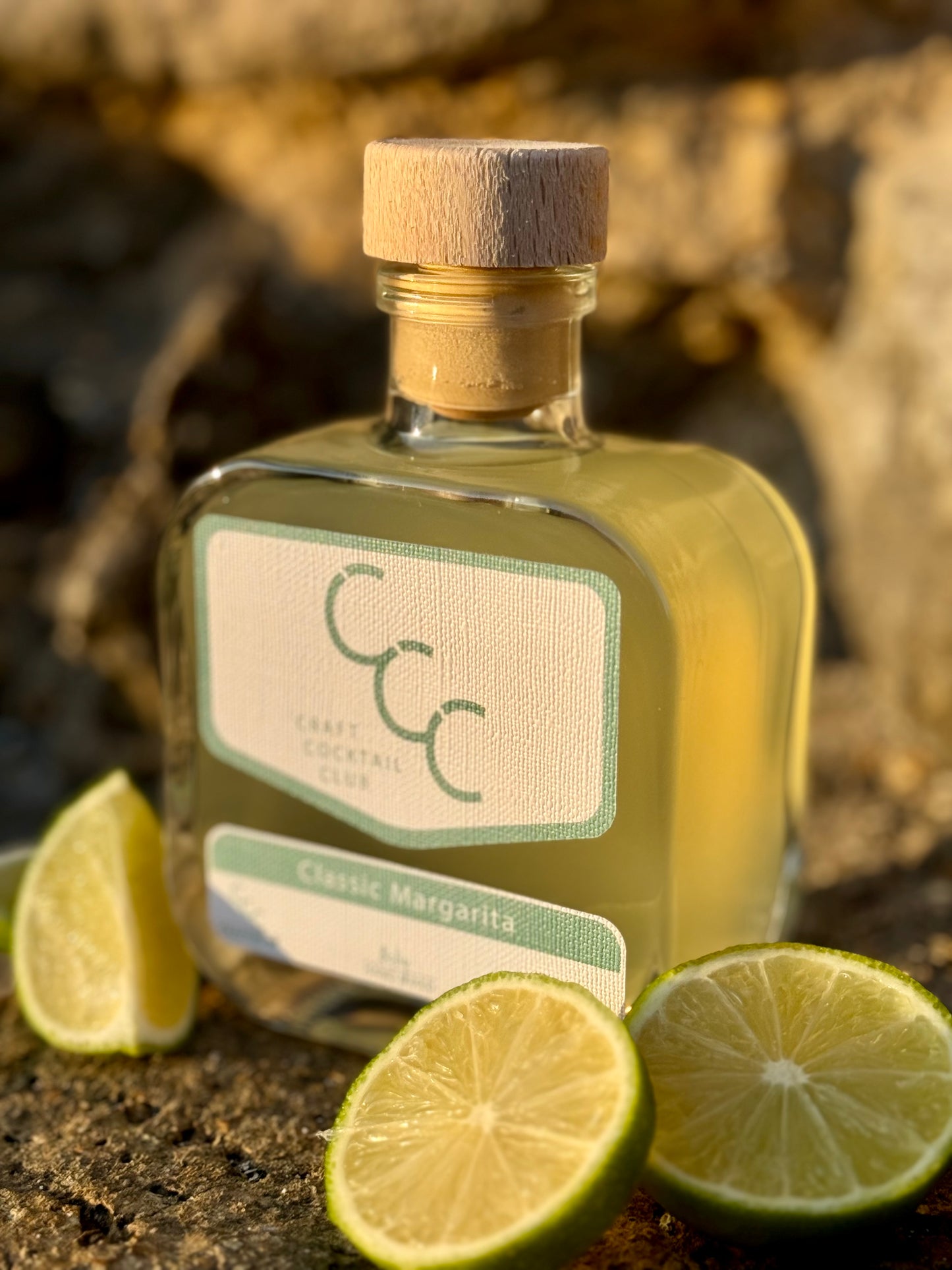 Picture of a square glass bottle of Craft Cocktail Club Margarita. It's a picture at sunset with rocks in the background, at the base of the bottle are cut fresh limes. the label reads Classic Margarita 