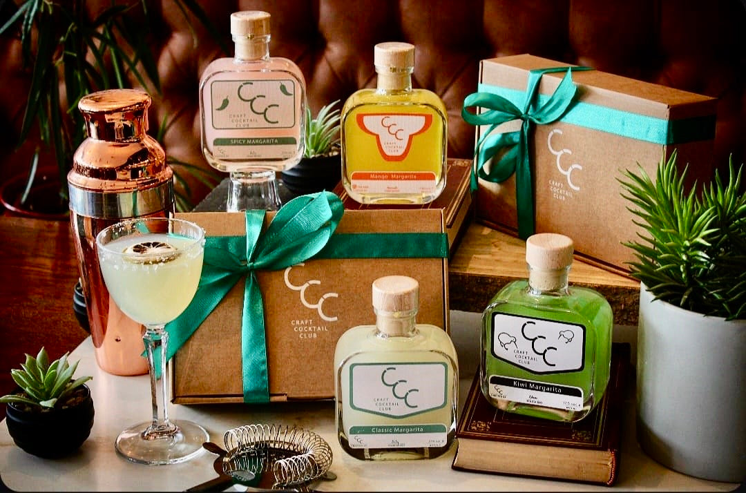 Craft  Cocktail Club bottles, a cocktail glass, and gift boxes on a wooden surface with plants.