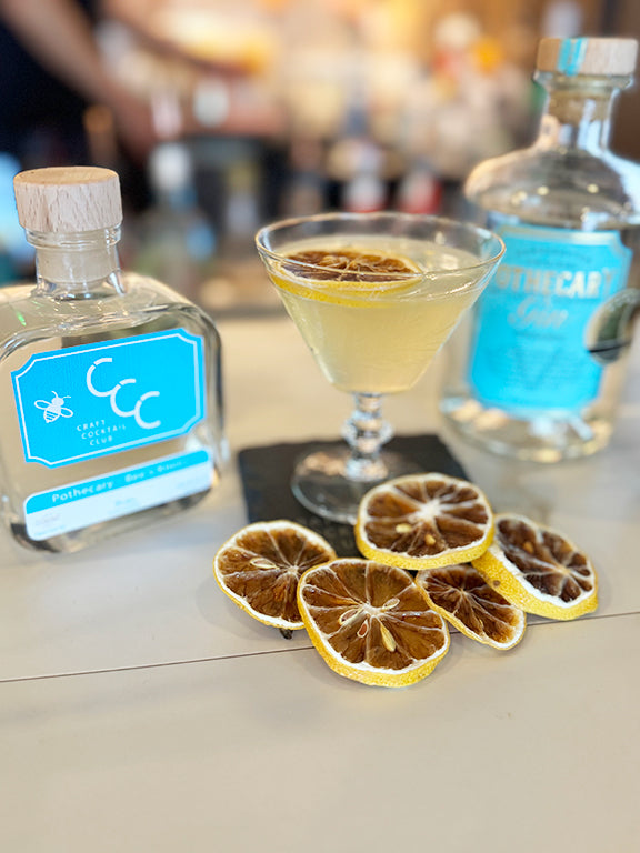 Picture  of a gin bottle next to a yellow cocktail with lemons around the base of the glass in a bar setting 