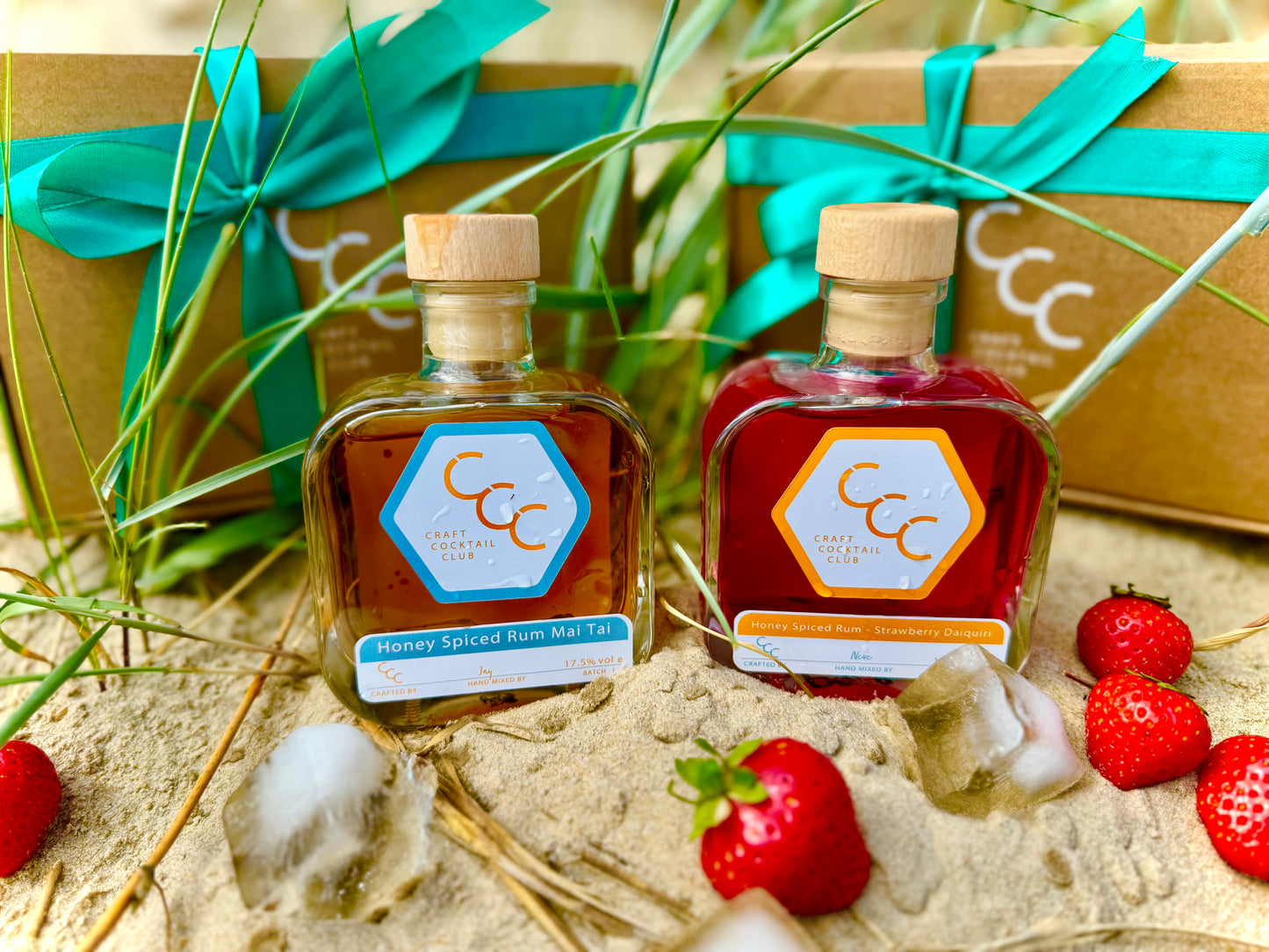 Picture of two Square Glass bottles of Pre mixed cocktails sat on a beach with a gift box in the background- The cocktails are Craft Cocktail Club Mai Tai and Strawberry 