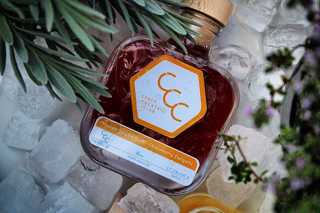 Picture a  bottles of ready to drink cocktails surrounded by ice cubes with foliage blurred on the edge of the picture, labels on the bottles say Craft Cocktail Club Strawberry Daiquiri