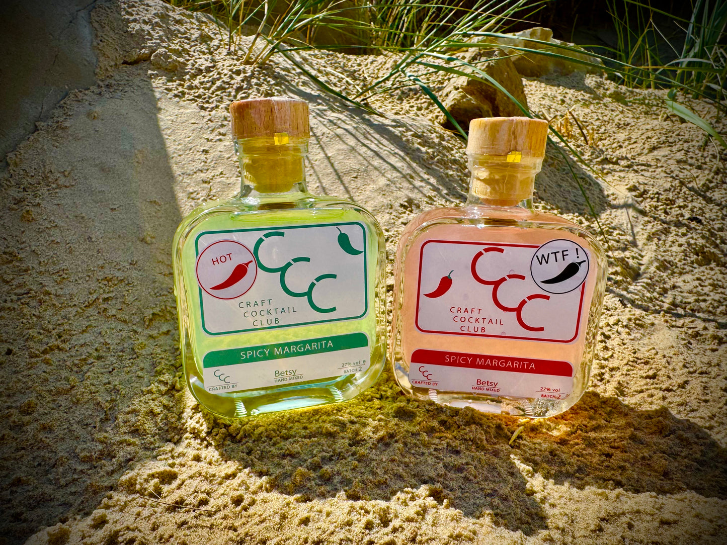 Two small bottles of spicy margarita with chili peppers on a beach, surrounded by fresh limes with a gift box in the background 