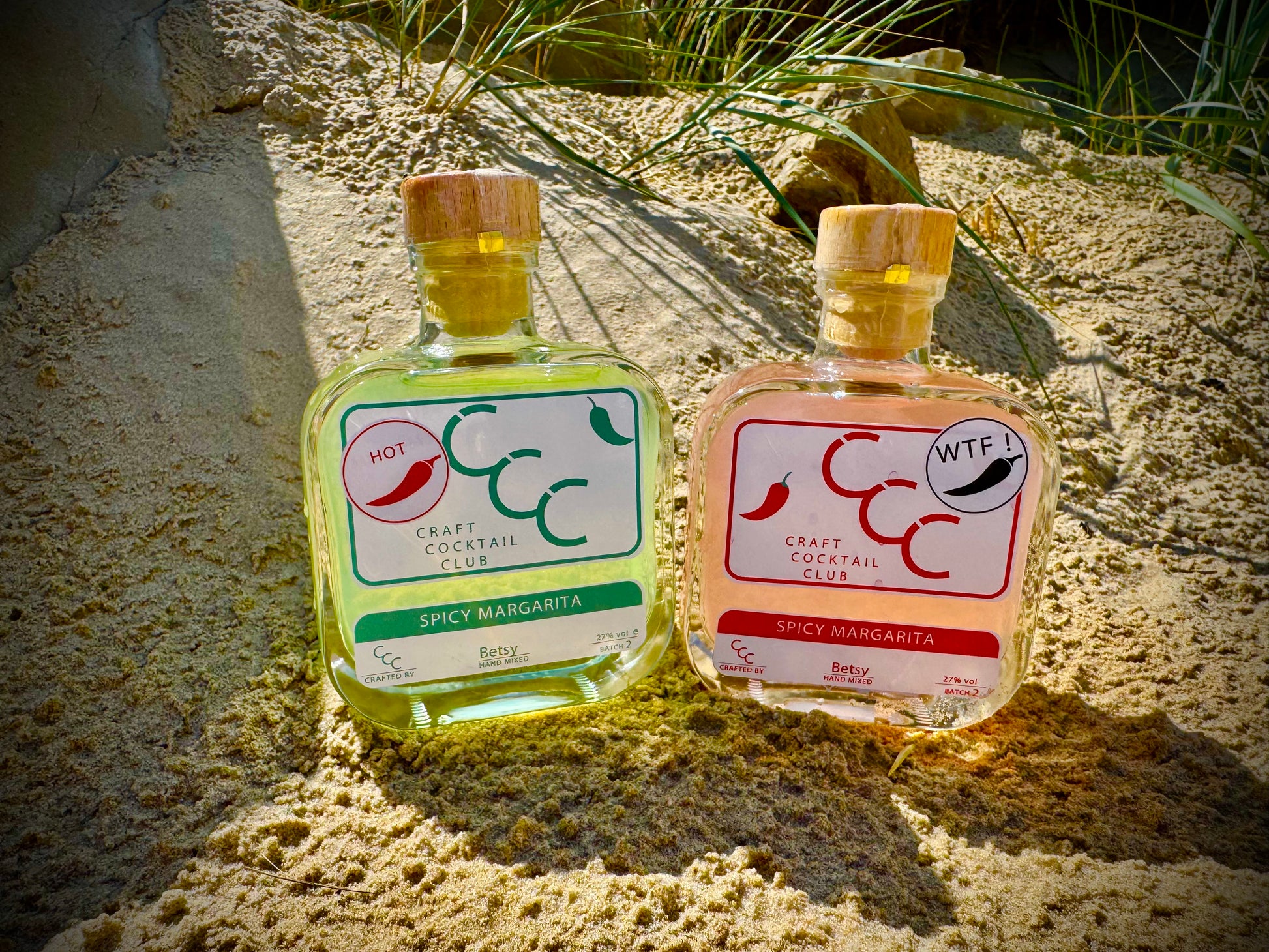 Two small bottles of spicy margarita with chili peppers on a beach, surrounded by fresh limes with a gift box in the background 