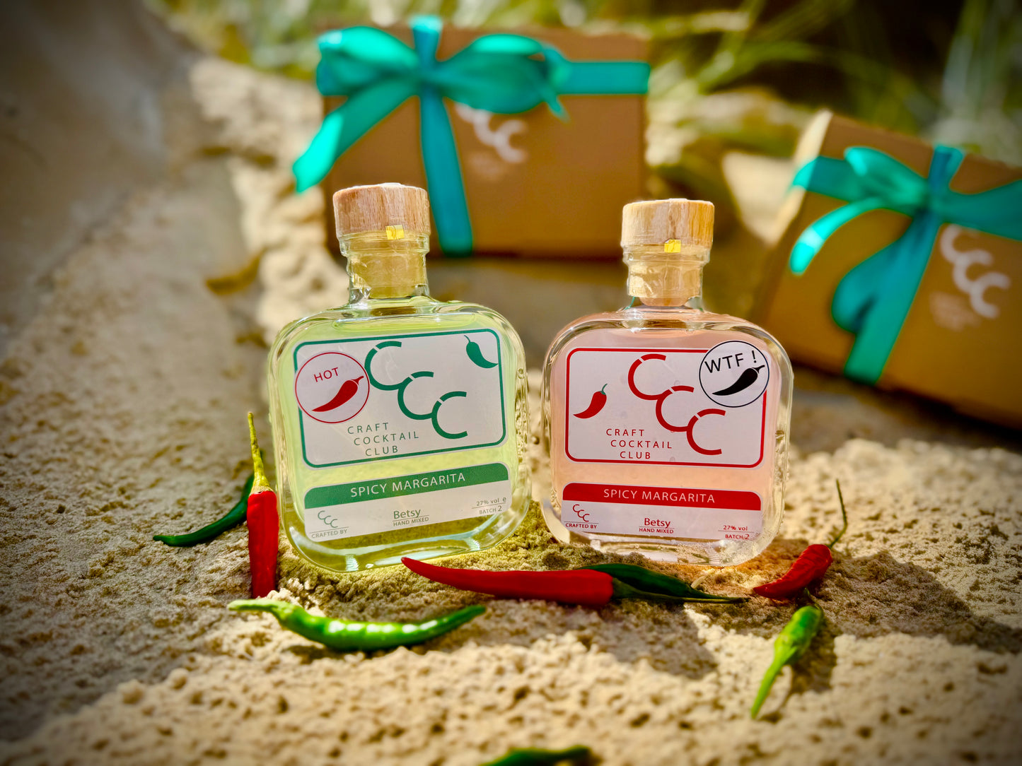 Two small bottles of spicy margarita with Chile peppers on a beach, surrounded by fresh limes with a gift box in the background 