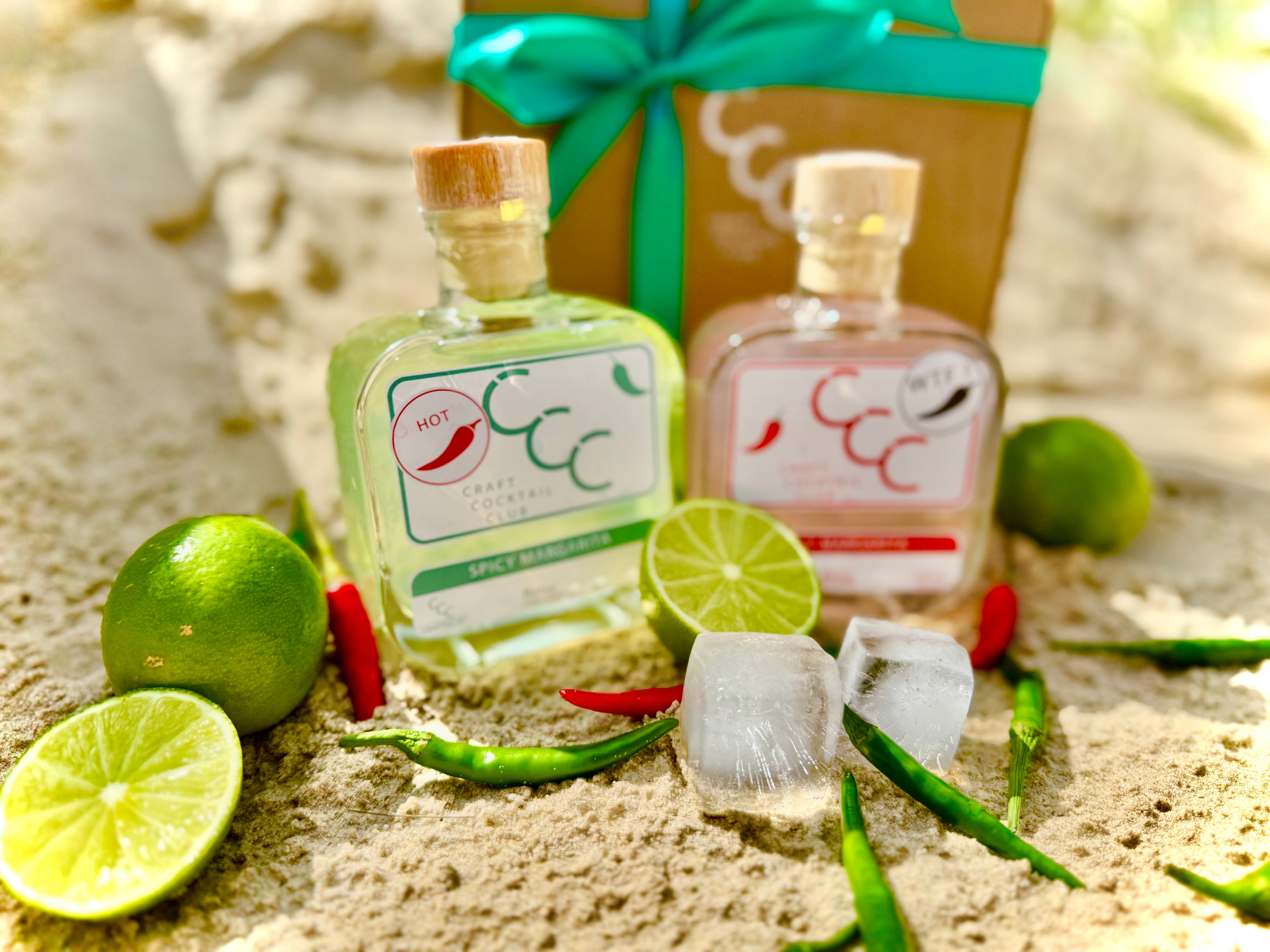 Two small bottles of spicy margarita with chillie peppers on a beach, surrounded by fresh limes with a gift box in the background - Label reads Craft Cocktail Club Spicy Margarita 