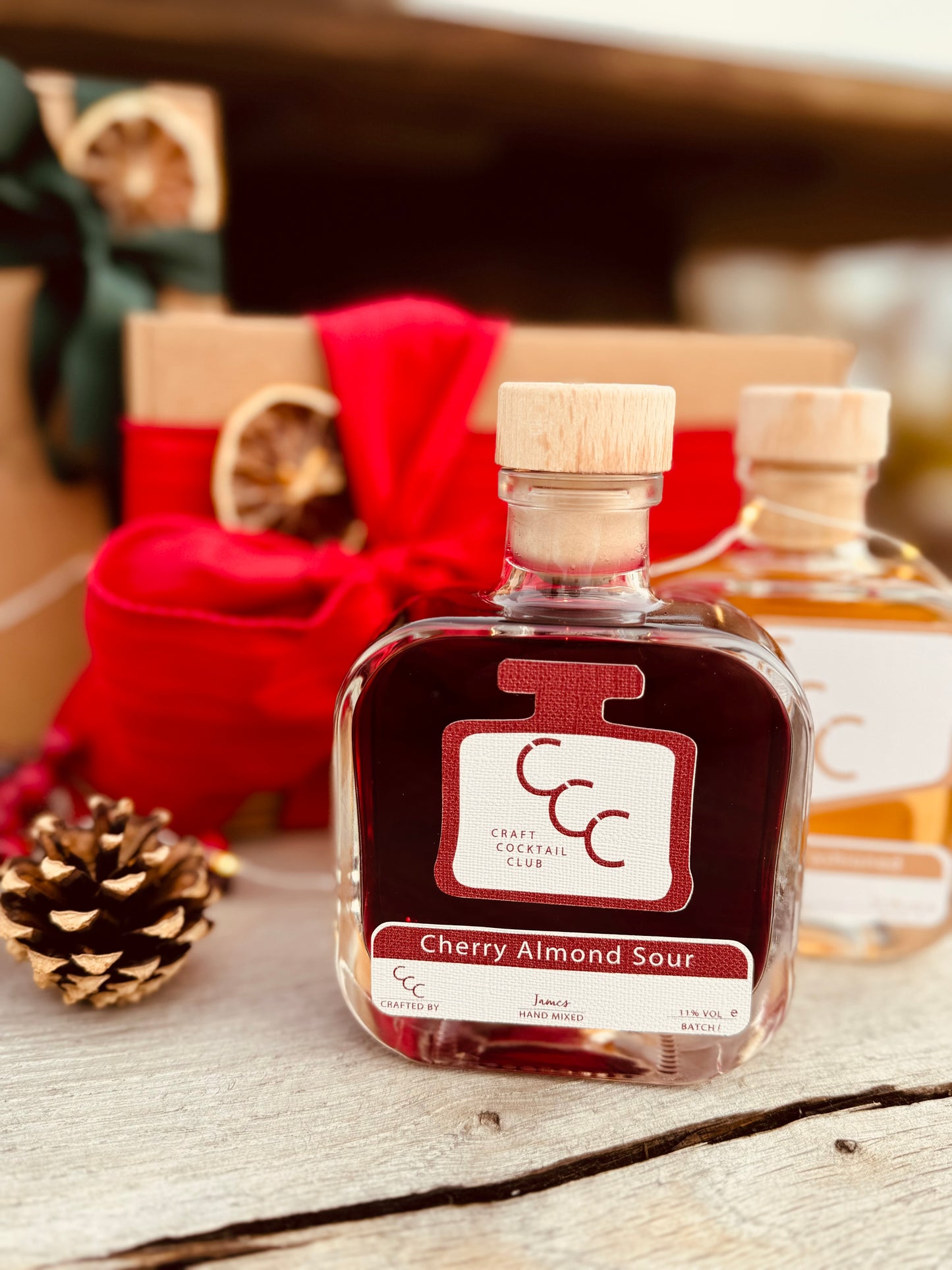Picture of a square glass bottle of craft cocktail club cocktail, with deep red liquid inside. It's surrounded by gift boxes with red and green ribbons with a pine cone to the side. The label reads cherry almond sour 