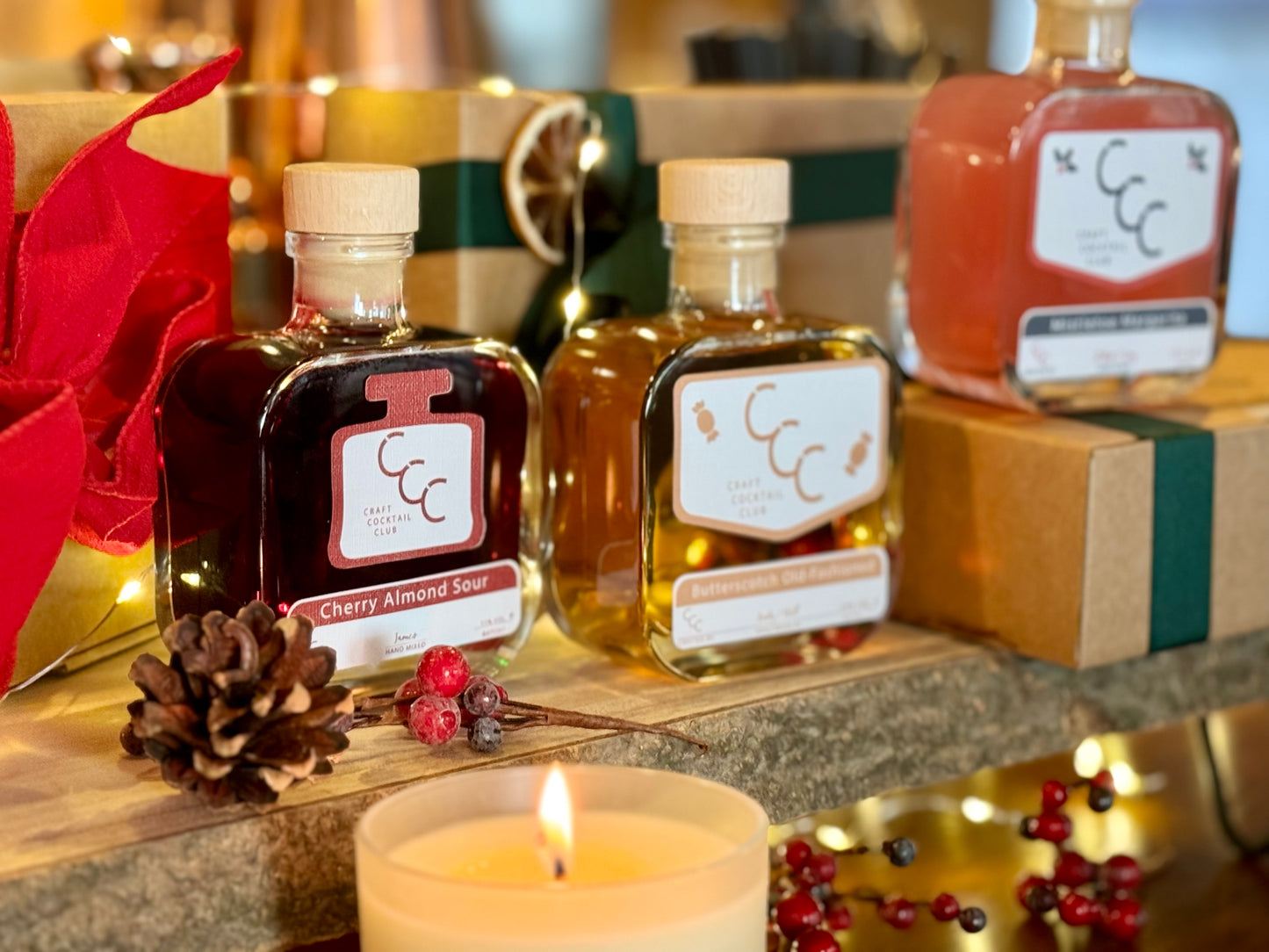A picture of there ready to drink cocktails in square glass bottles. One reads almond sour. they are sat on a rough wood board, with gift boxes and lights in the background, with a candle in the first surrounded by winter berries