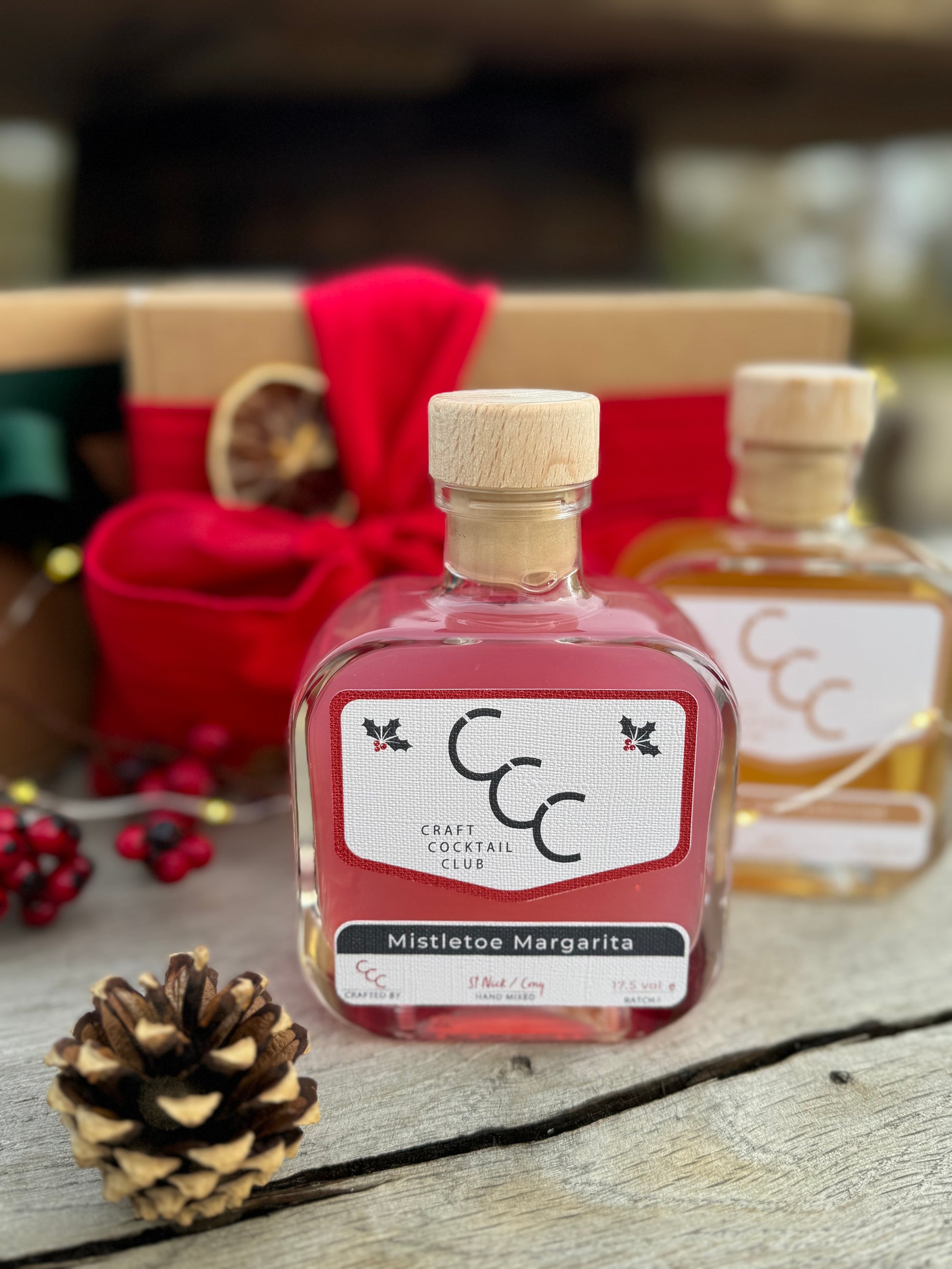 A picture of a Square glass cocktail bottle, sat on a wood table with a gift box in the background slightly blurred with a red ribbon. 