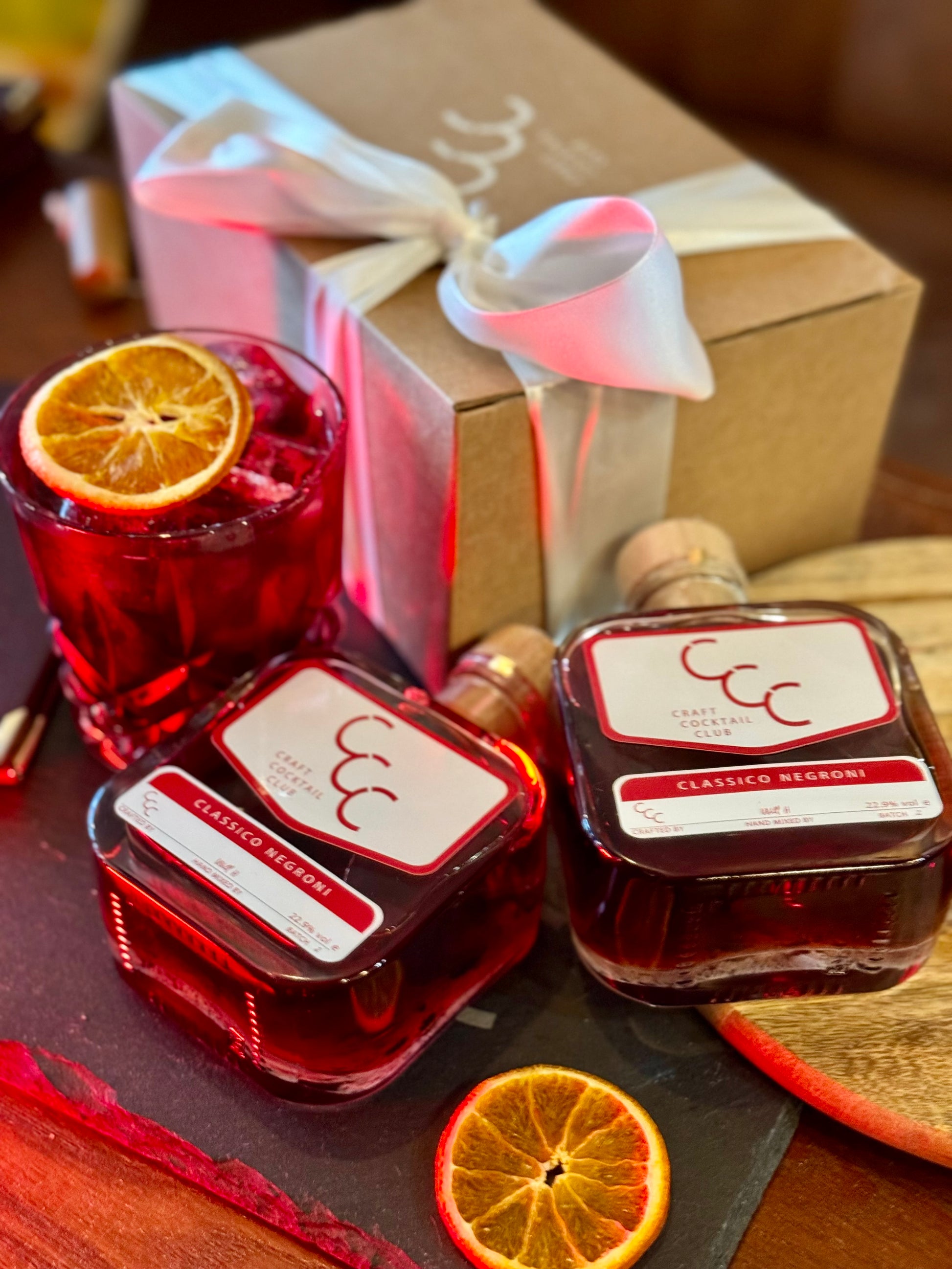 Picture of a gift box on a wooded table, next to the gift box is a small glass filled with a ruby red liquid, ice and a slice of orange on top, Next to the glass are two square glass bottles of Craft Cocktail Club Cocktails, the label reads Clasic negroni 