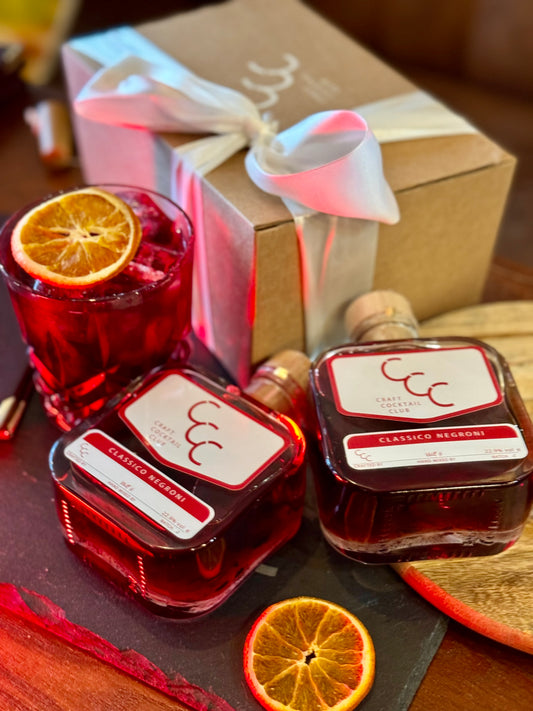 Picture of a gift box on a wooded table, next to the gift box is a small glass filled with a ruby red liquid, ice and a slice of orange on top, Next to the glass are two square glass bottles of Craft Cocktail Club Cocktails, the label reads Clasic negroni 