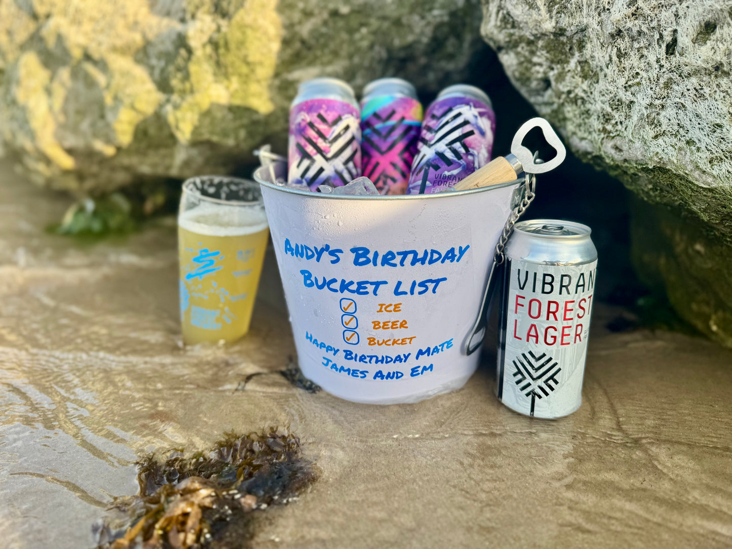 Picture of a personalised bucket of beers on a beach, the table is personalised with a personal birthday list and the persons name (In this case Any's birthday Bucket). The bucket is full of beers from Vibrant Forest Brewery 