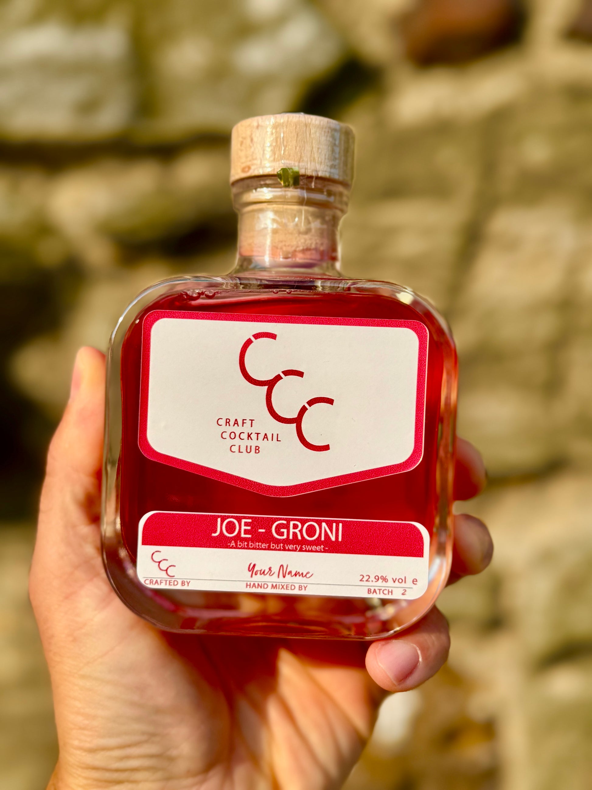 Bottle of a personalised  Craft Cocktail Club bottle filled with ruby red liquid held in someone's hand against a stone background,  the label reads  Joe Groni 