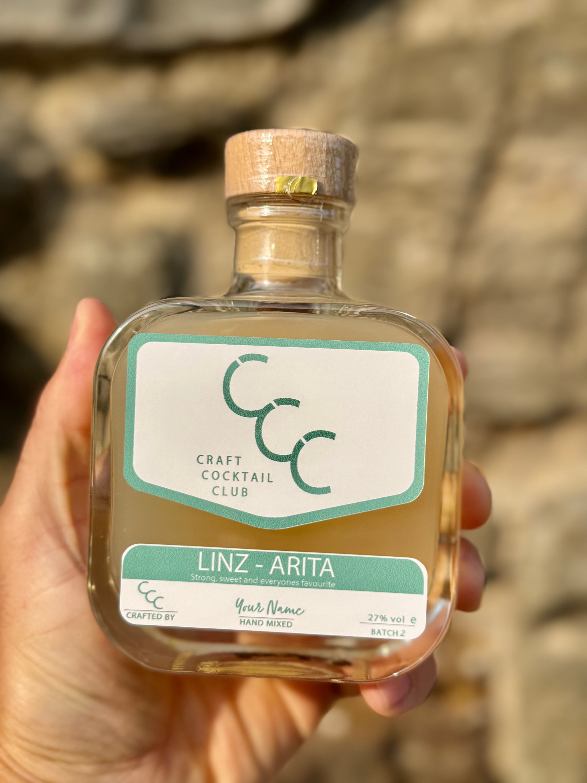 Bottle of Craft Cocktail Club Margarita with a personalised label held by a hand against a stone wall background