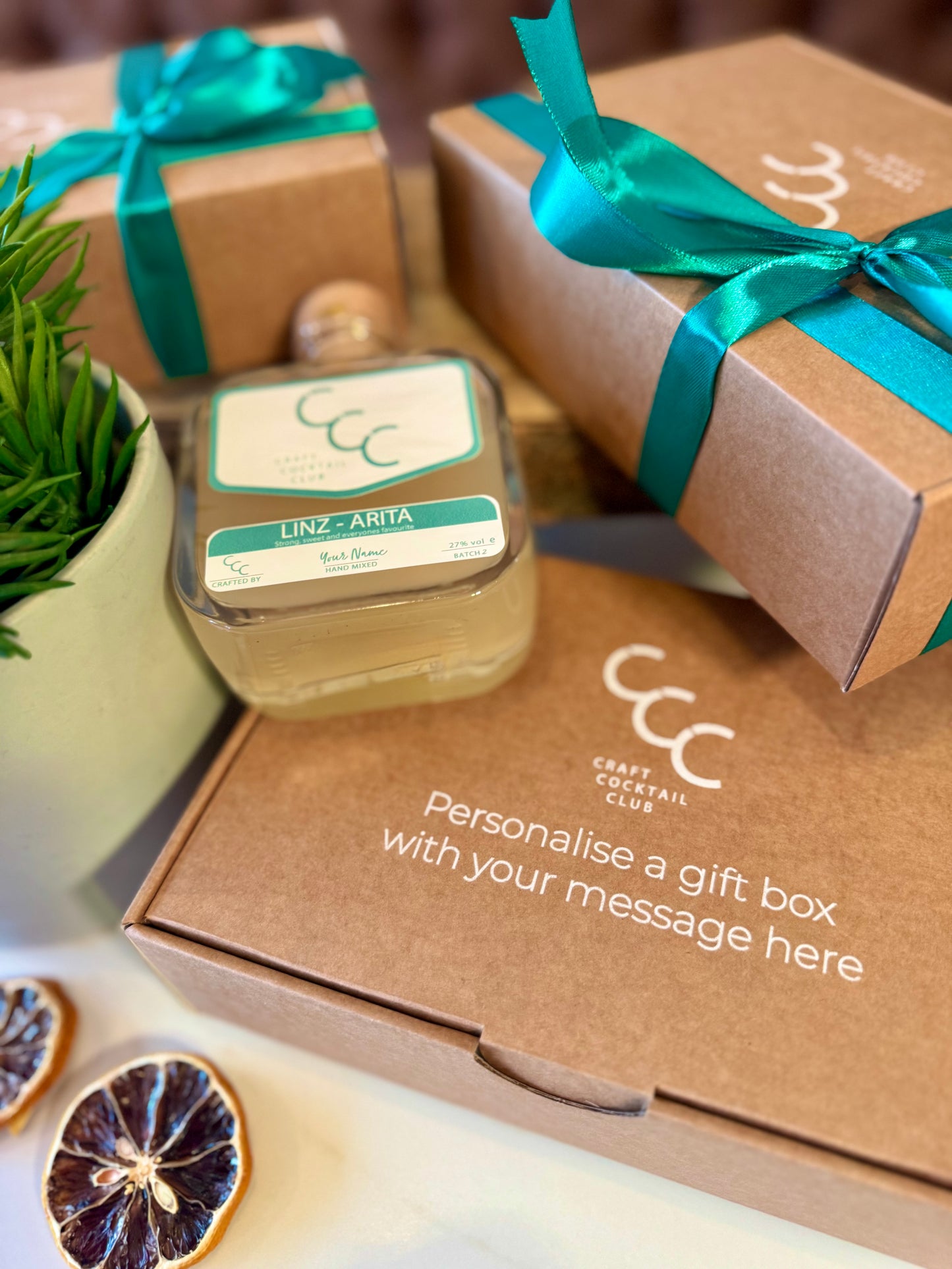 Bottle of a personalised  Craft Cocktail Club bottle filled with lime green  . The Square glass bottle is surrounded with gift boxes with ribbons on the gift box ir reads - Personalised gift box with your message here 