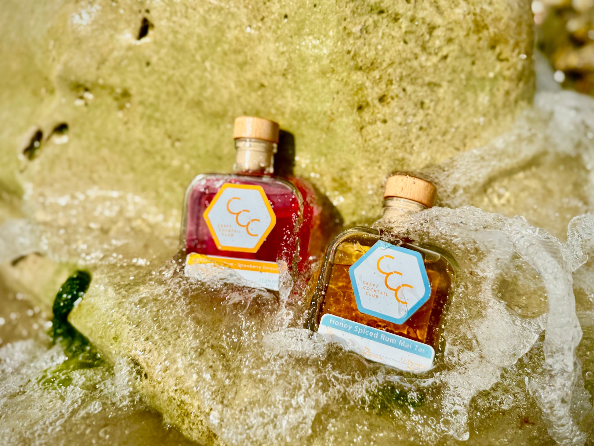 Picture of two bottles of Craft Cocktail Club ready to drink cocktails on a rock with the sea splashing over them on a sunny day. The cocktails Are a mai star and strawberry daiquiri
